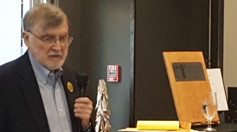 A man in a suit speaking into a microphone in a conference room or meeting space.