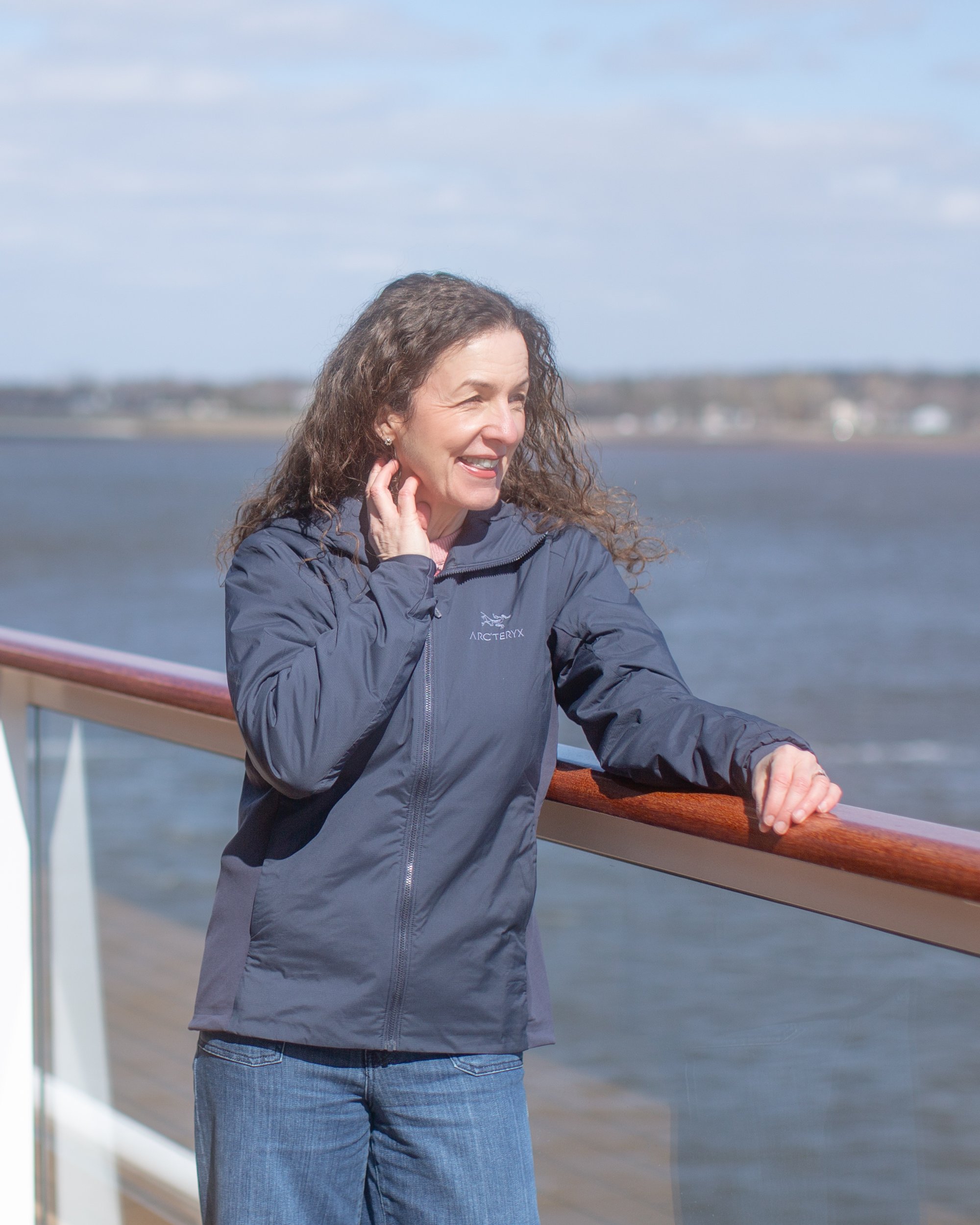 Rona Wheeldon -A woman with curly brown hair wearing a navy blue Arc'teryx jacket and blue jeans, smiling. She is on an expedition ship with a body of water and a distant shoreline in the background.