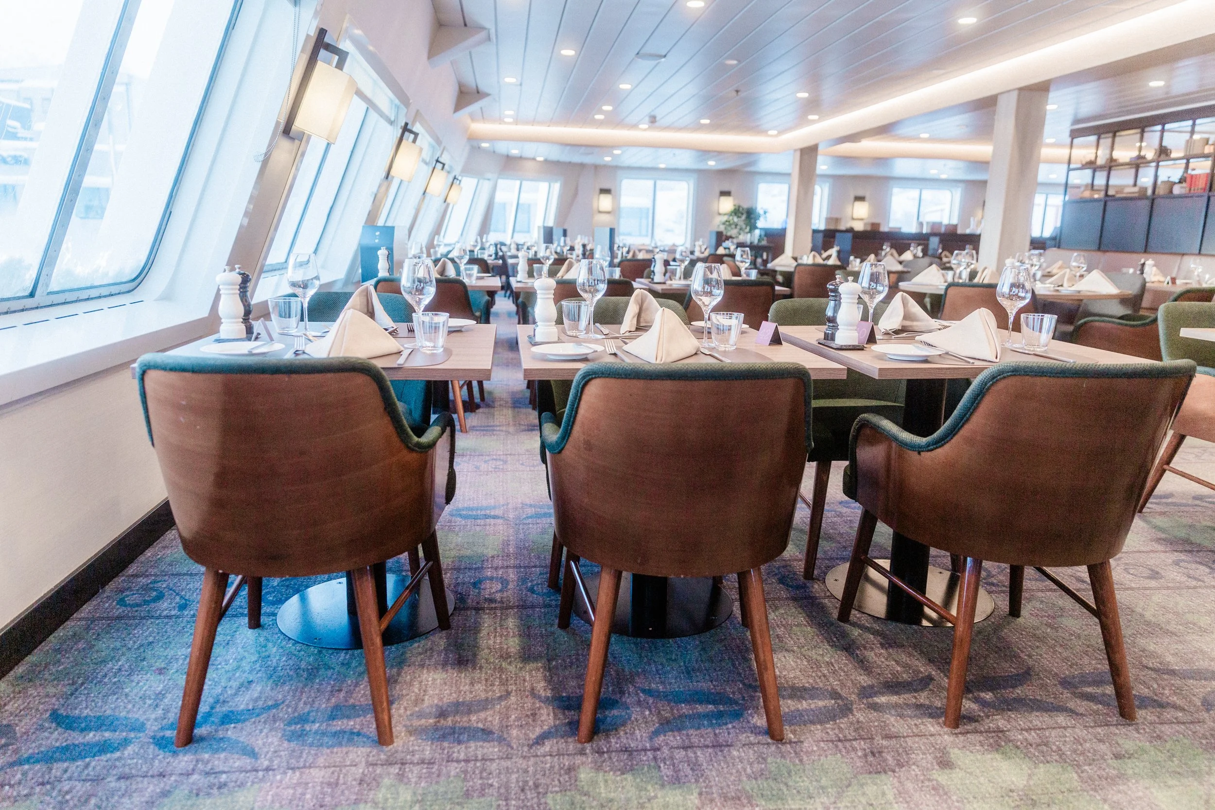 Dining area of Aune Restaurant on MS Spitsbergen, with neatly set tables, blue upholstered chairs and large slanted windows letting in natural light.