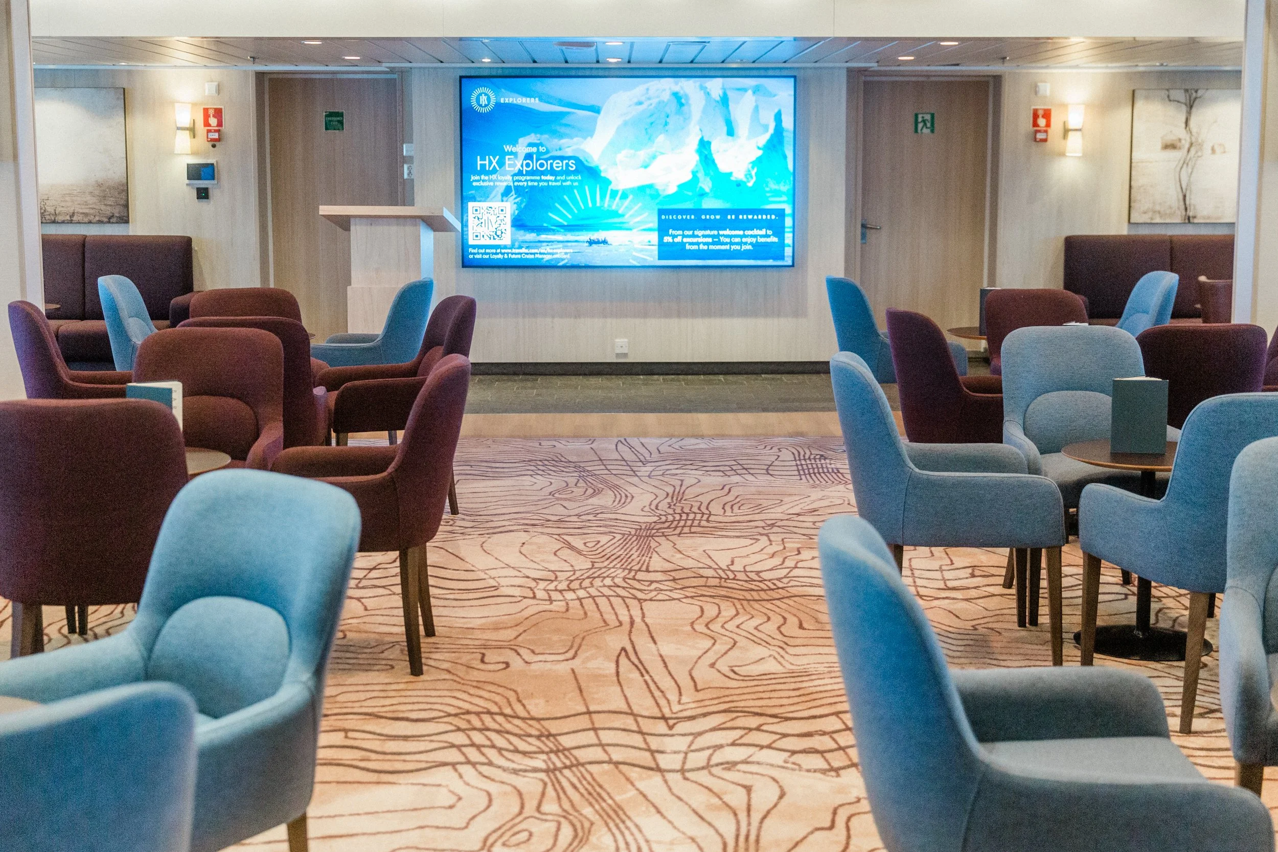 Lounge area on MS Spitsbergen with blue and brown chairs, small round tables, a patterned carpet and a large screen at the front displaying an HX Explorers welcome message.