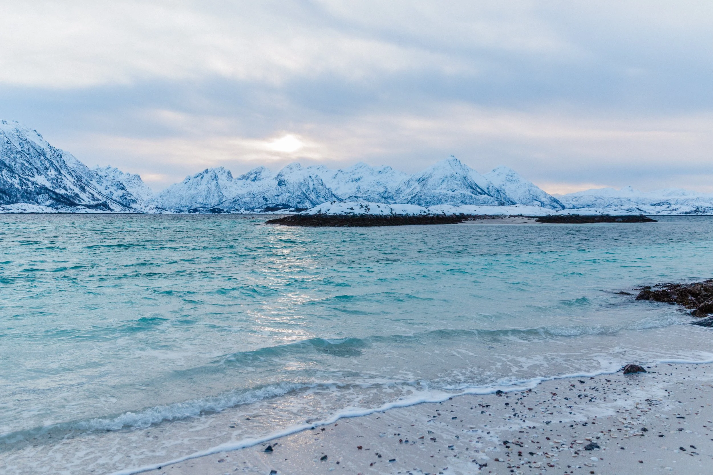 Snow-capped mountains in the background with a body of water in the foreground and a rocky, sandy beach in Norway.
