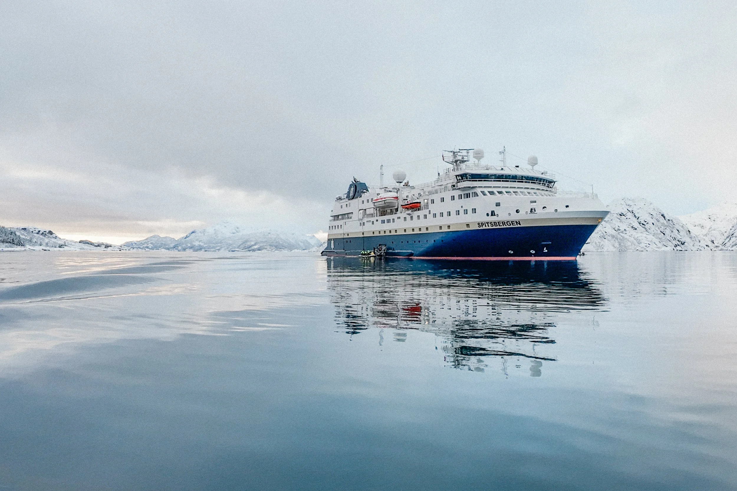 An expedition cruise ship named MS Spitsbergen floating on calm icy waters, with snow-covered mountains in the background under a cloudy sky.