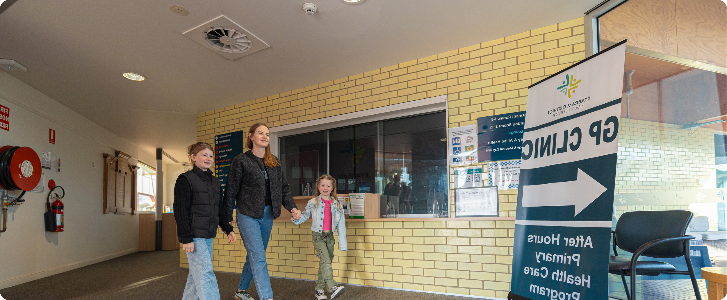 A woman and two young girls walking into a health center, holding hands, with a sign indicating directions for the GP clinic and other healthcare services, inside a reception area with yellow brick walls and large windows.