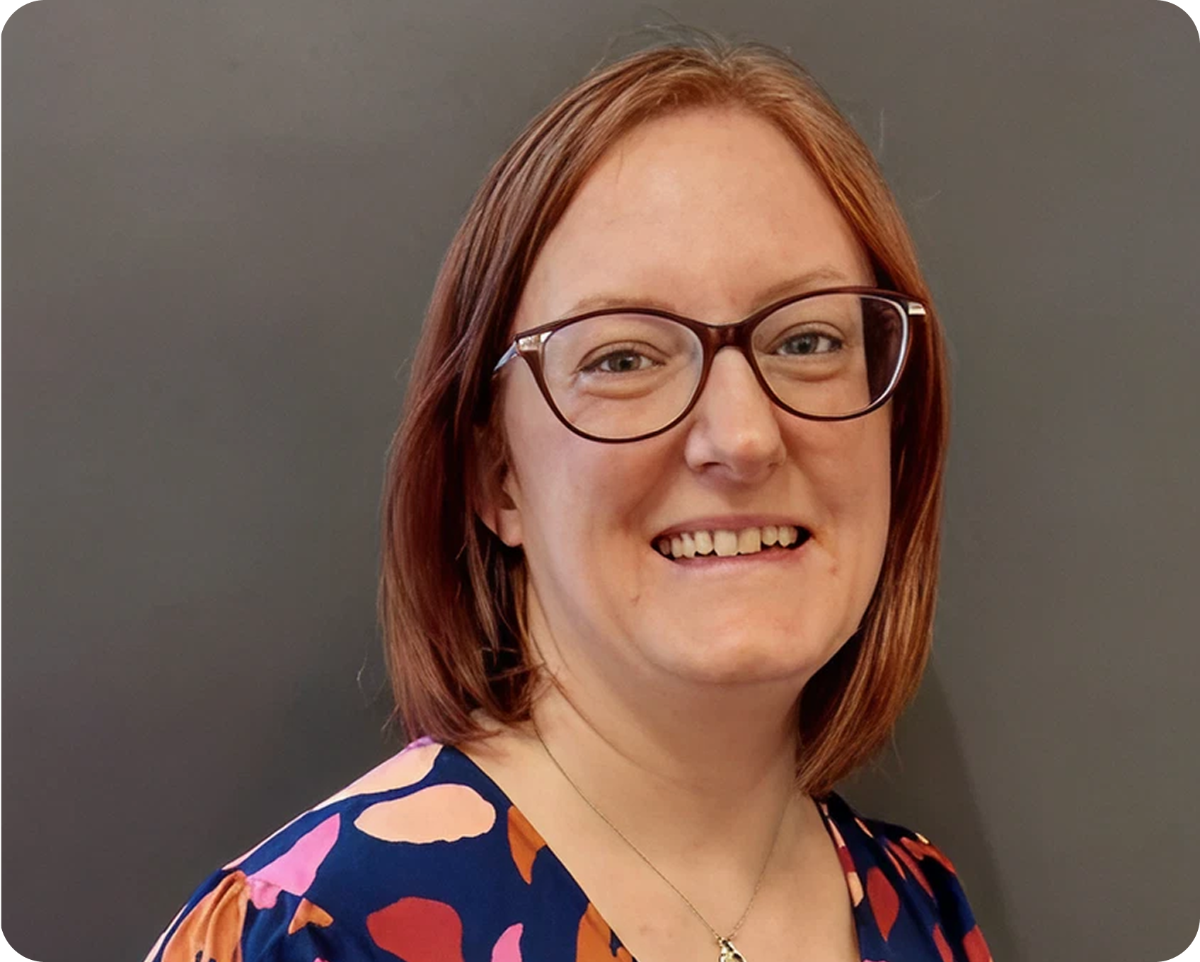 A smiling woman with red hair, glasses, and a colorful abstract-patterned top stands against a dark gray background.