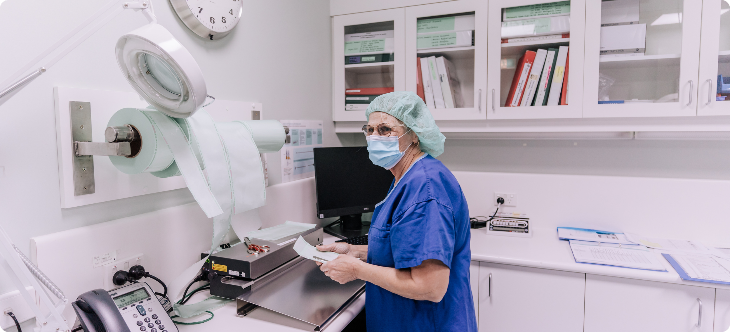 A healthcare worker in blue scrubs, wearing a surgical mask, glasses, and a hair cap, is working in a hospital or medical lab with medical equipment and shelves of binders in the background.