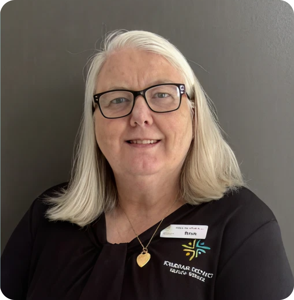 A woman with long, blonde hair, glasses, and a friendly smile, wearing a black top with a colorful logo and a name tag, against a plain gray wall.