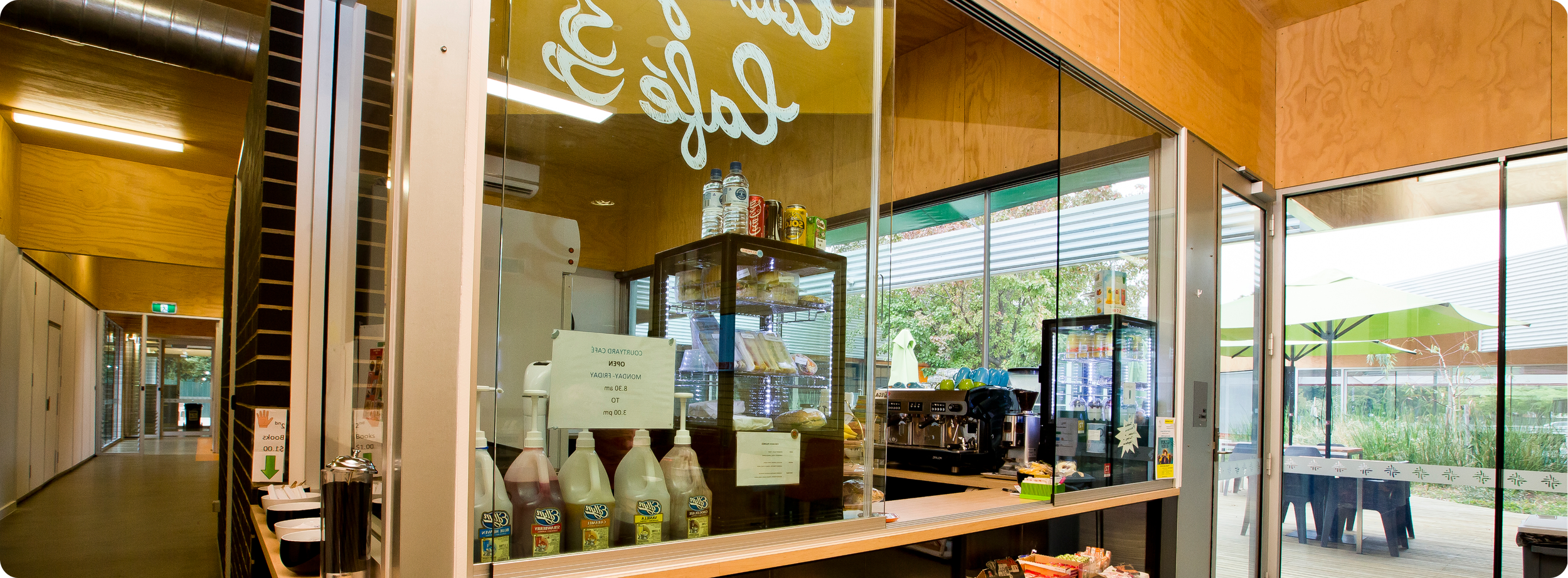 Inside a modern cafe with a wooden interior, a counter with a glass display case containing baked goods, bottles of water, and cans of soda on top. There is a coffee machine and a small refrigerator with drinks. Large glass windows and doors reveal outdoor seating with green umbrellas and black chairs.