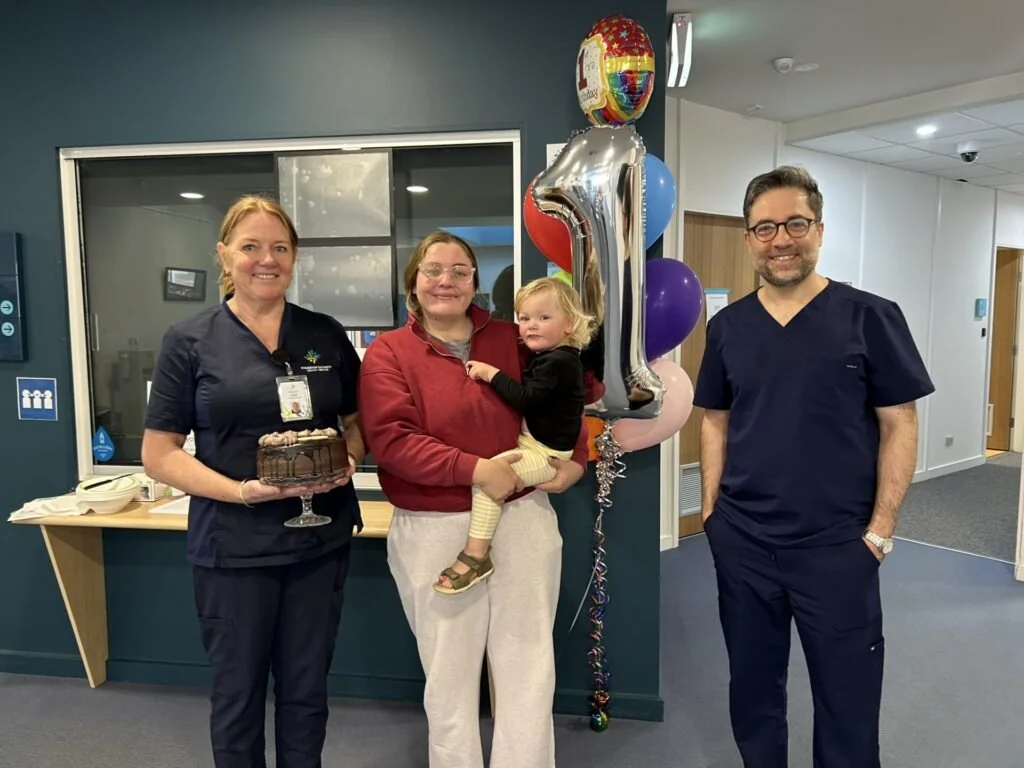 People celebrating a child's first year at a healthcare facility, with balloons, a cake, and a nurse.