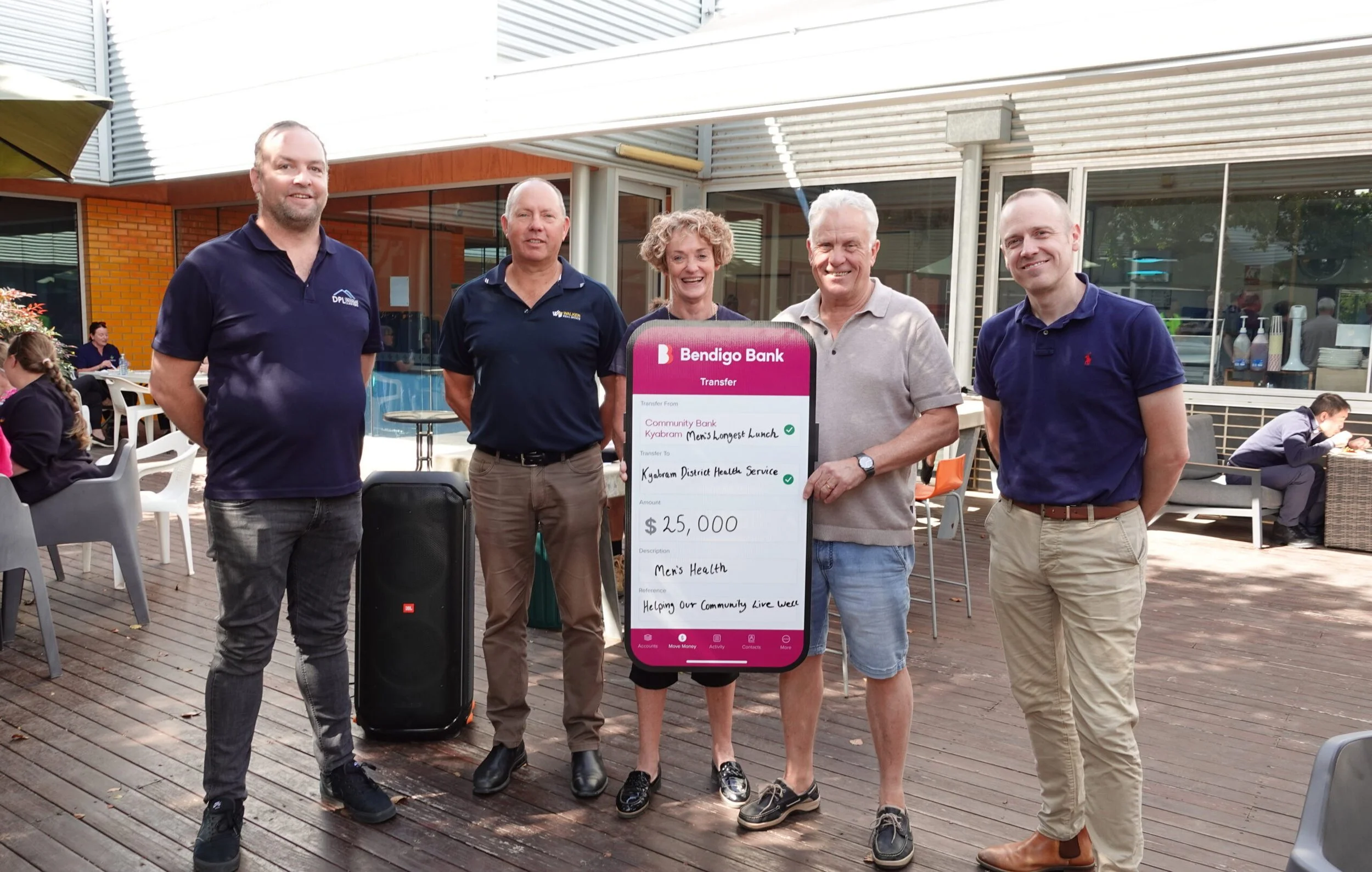 Group of five people smiling and standing outdoors, holding a large mock check for $25,000 from Bendigo Bank, with a modern building and other people seated at tables in the background.