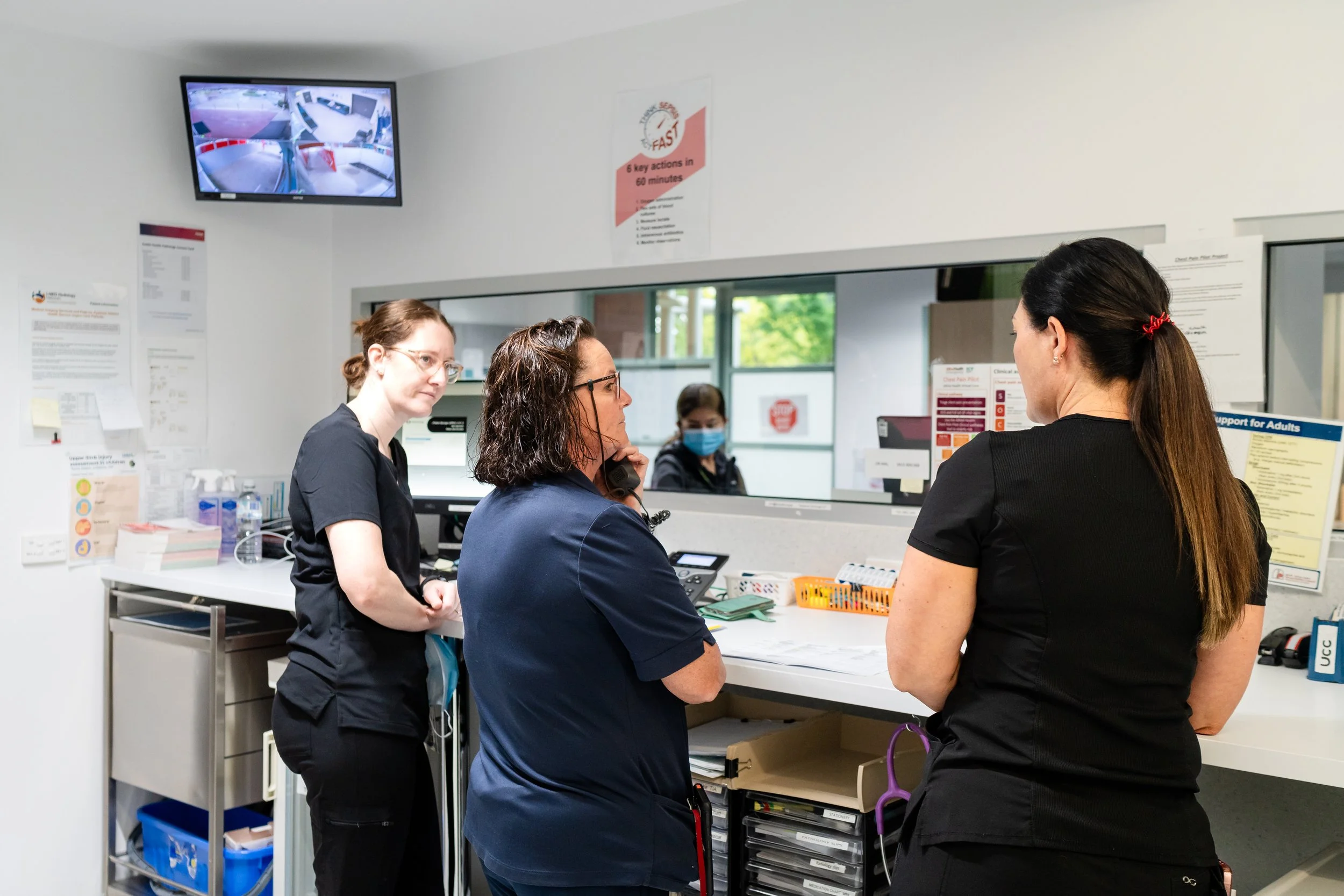 Three women and one woman in the background with a mask are in a hospital or clinic registration or reception area. They are engaged in conversation, with one woman speaking to another, possibly about check-in or medical matters. The setting features a counter, various informational posters, a monitor displaying surveillance footage, and medical supplies.