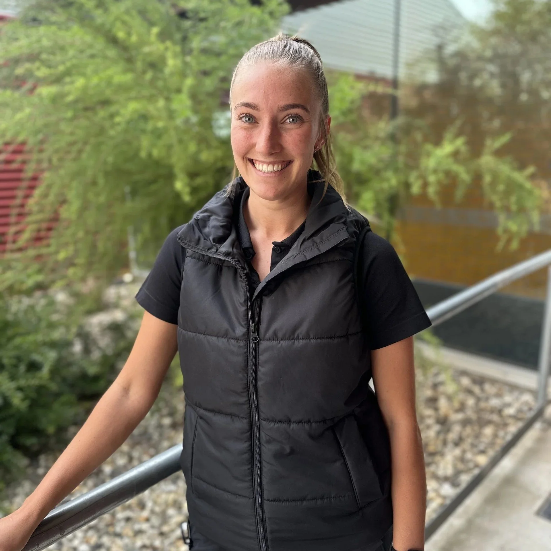 Close-up of a smiling young woman with blonde hair tied back, wearing a black sleeveless puffer vest, outdoors with green trees and a building in the background.