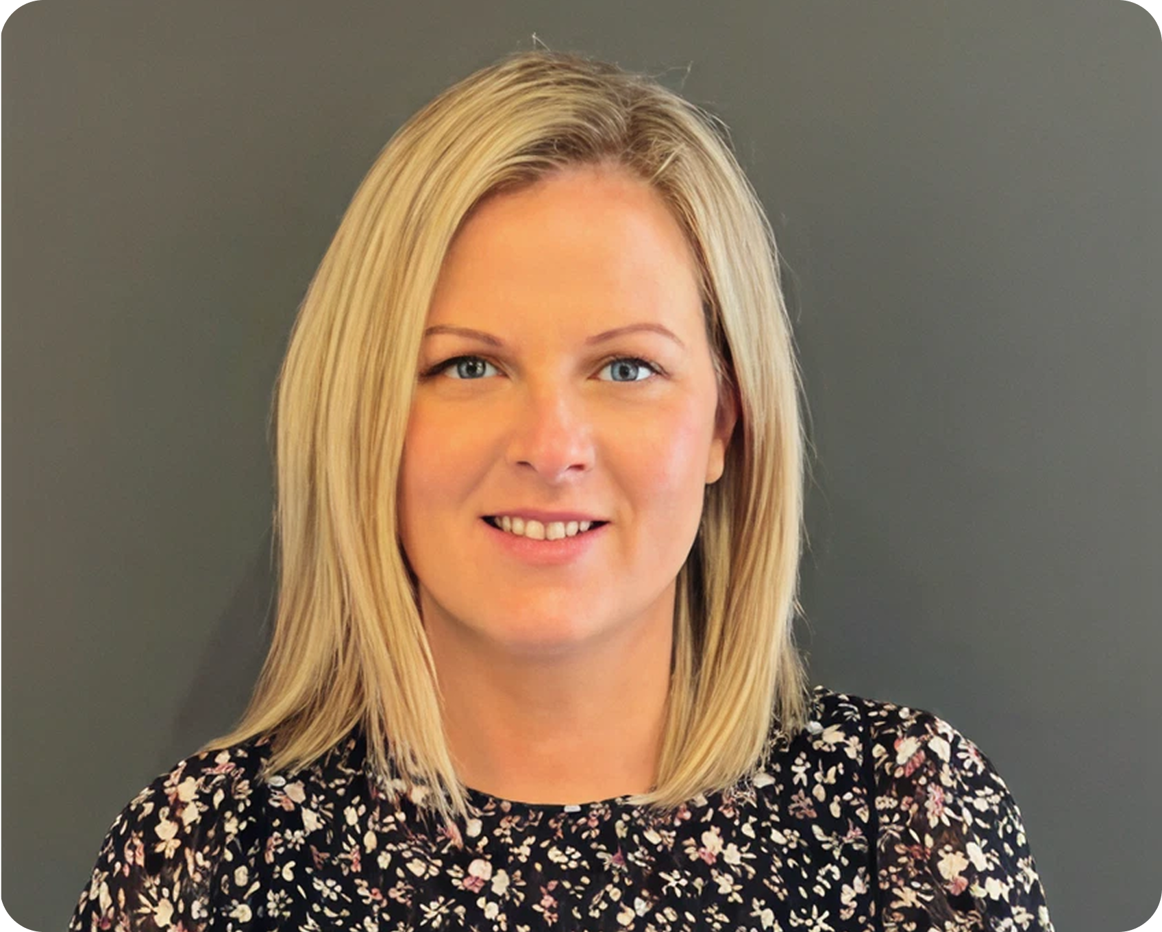A woman with blonde hair wearing a black floral shirt, smiling against a gray background.