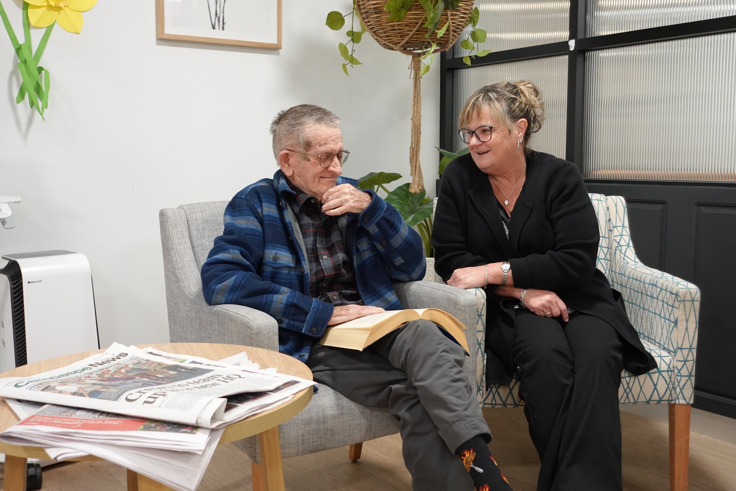 An elderly man with glasses wearing a blue plaid jacket and a woman with glasses and earrings are sitting in a cozy indoor space decorated with plants and artwork, smiling and engaging in conversation. There are newspapers on a small table in front of them.