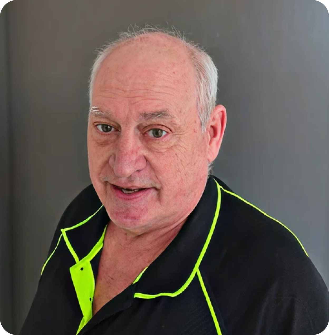 Close-up photo of an older man with short gray hair, wearing a black shirt with neon yellow piping, standing against a dark gray background.