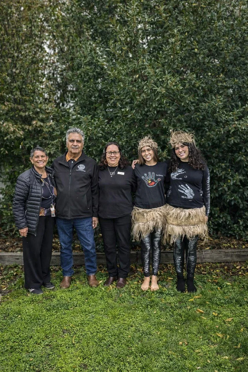 Group of five people standing outdoors on grass, with large green bushes in the background. Two women are dressed in traditional or cultural attire with straw skirts and headpieces. The others are dressed casually or professionally, smiling for the photo.