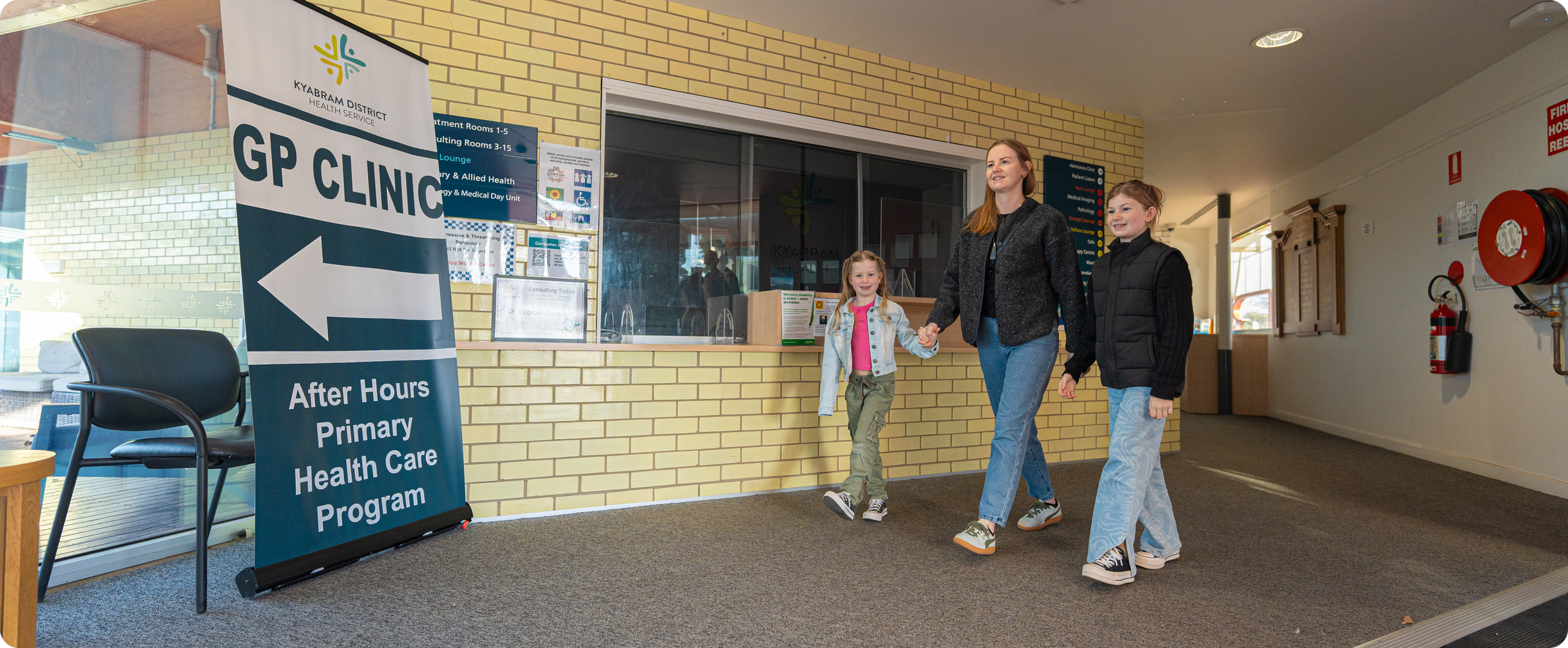 A woman with two young girls walking hand in hand inside a healthcare facility, with a sign indicating the GP Clinic and primary healthcare services nearby.
