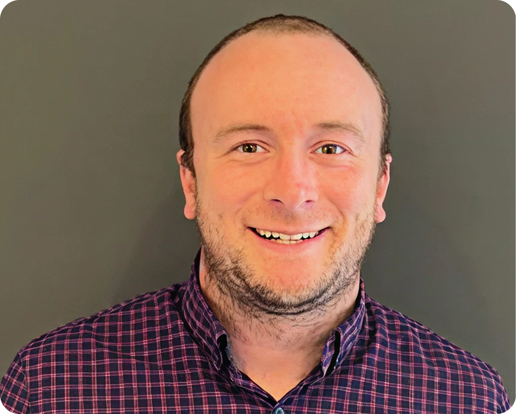 A smiling man with short hair, light skin, and a beard, wearing a dark purple checkered shirt, standing against a plain gray background.