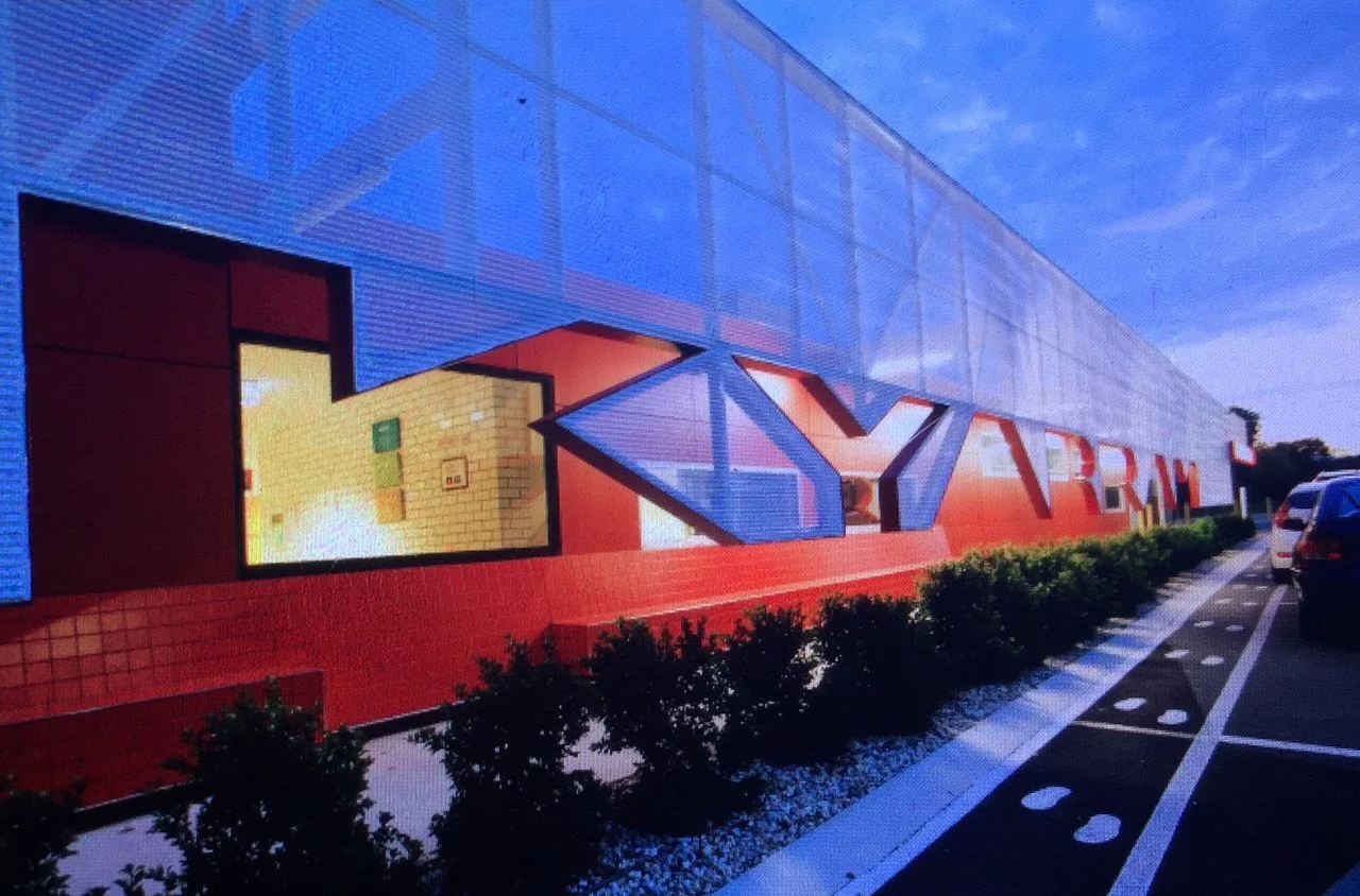 Exterior view of a modern building with large glass panels and bright red accents, with a row of green bushes in front and parked cars on the side.