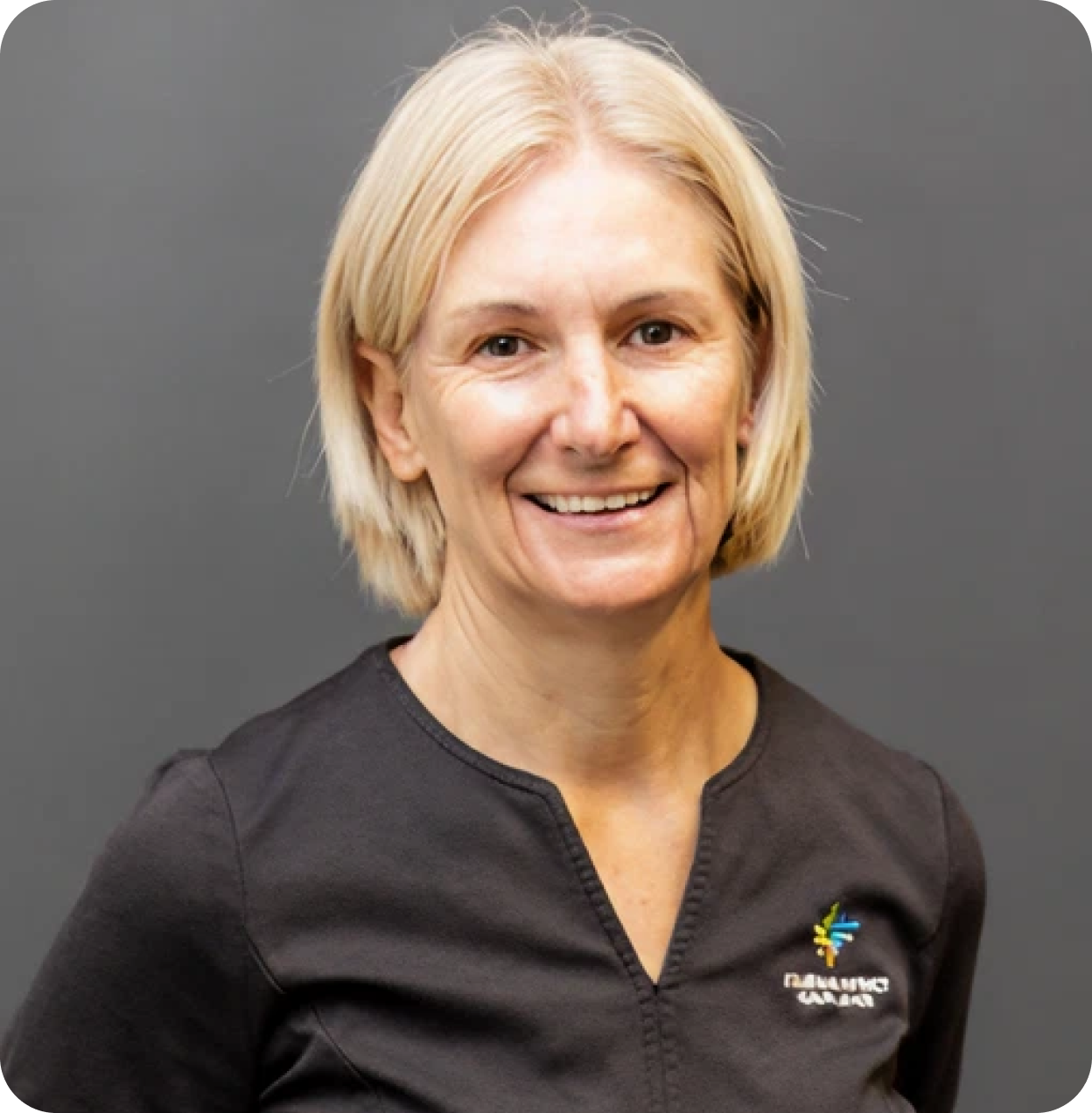 A smiling woman with blonde hair, wearing a black top with a small logo on the chest, standing against a gray background.