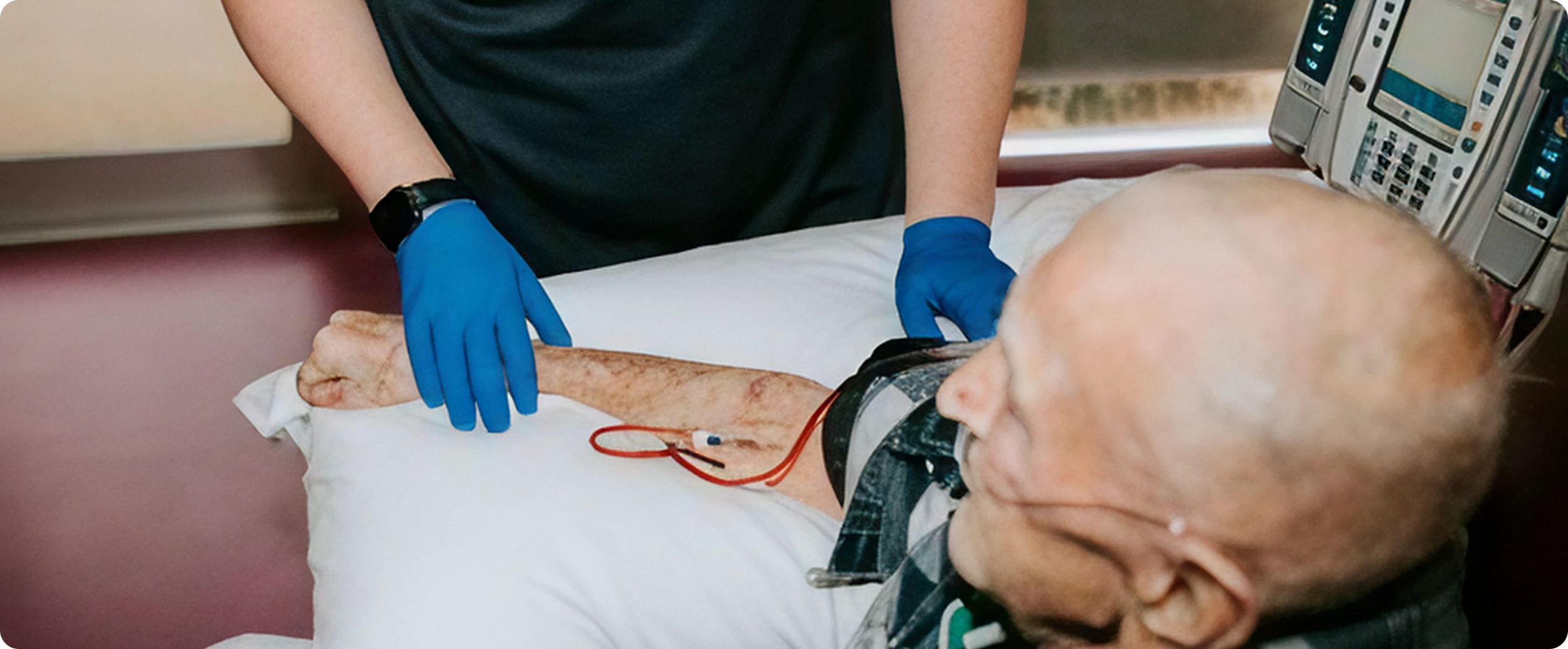 A healthcare worker with blue gloves caring for an elderly patient lying in a hospital bed, with medical monitors in the background.