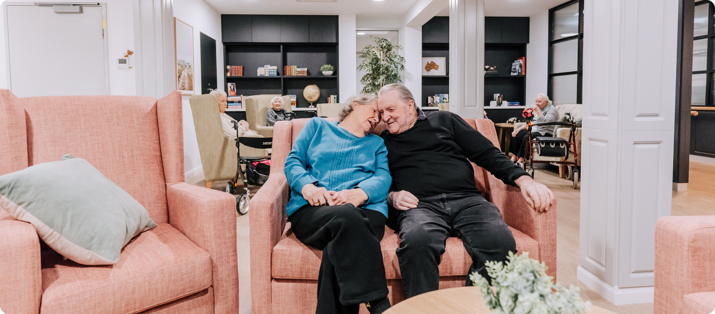 Two elderly people sitting closely on a pink sofa, sharing a tender moment with their foreheads touching. They are in a communal space with other elderly individuals in the background, some using walkers, and behind them are black bookshelves and a large window.