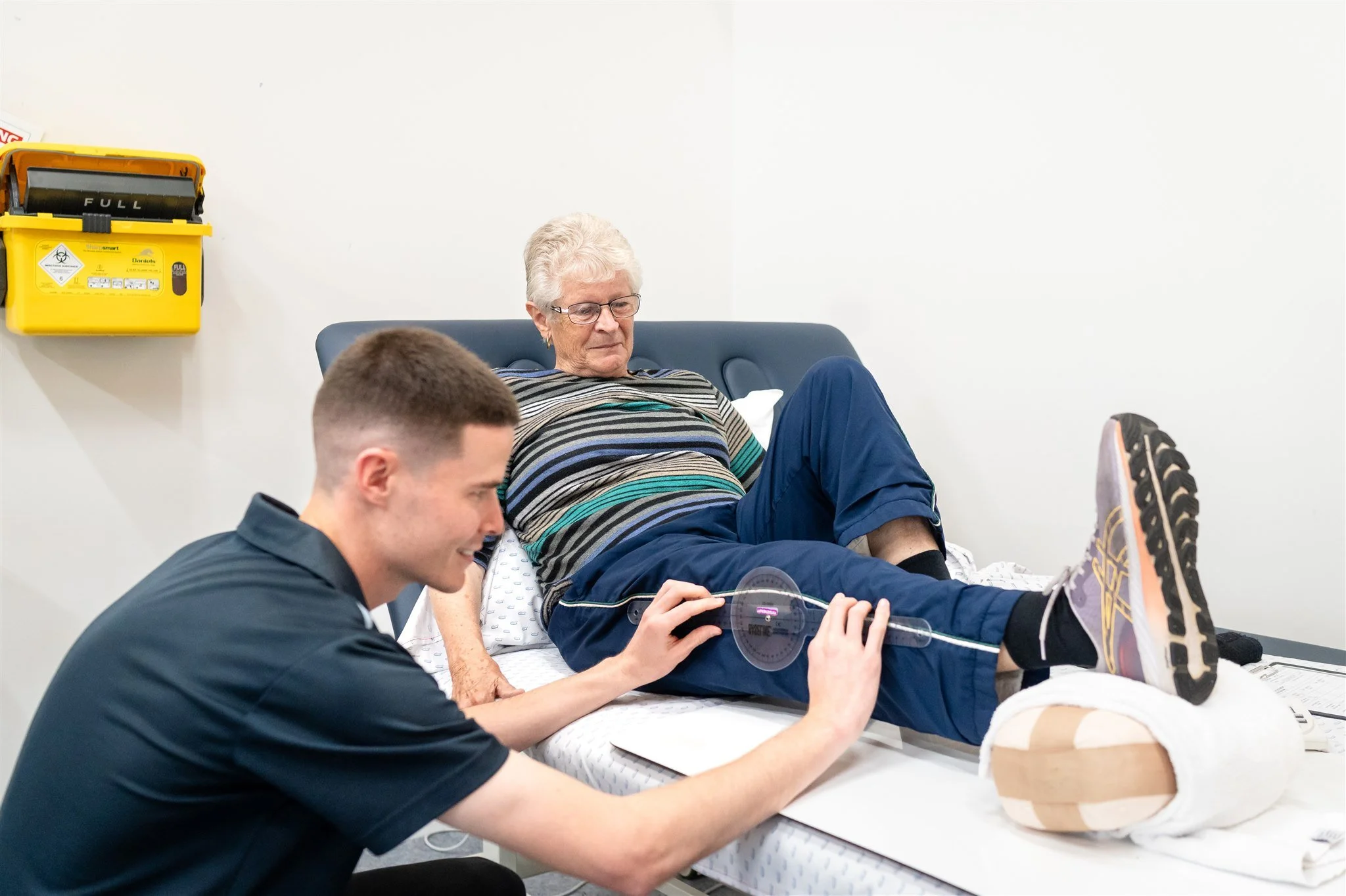 An elderly woman with white hair, glasses, and wearing a striped shirt, sits on a medical examination table with her feet up, while a young male medical professional in black scrubs checks her leg using a protractor-like tool. A medical dummy or mannequin is on the table beside her. The scene is set in a clinical room with a yellow biohazard sharps container on the wall.