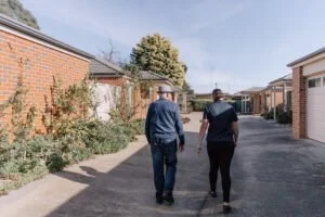 Two people, a man and a woman, walking down a residential alley with brick houses on each side and a tree in the background.