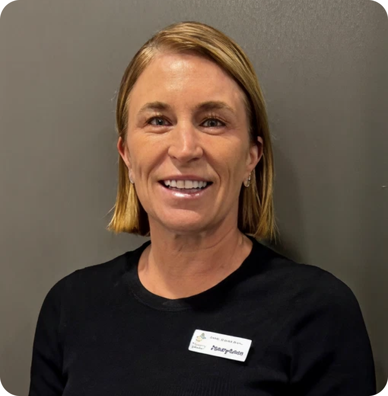 Portrait of a smiling woman with shoulder-length blonde hair, wearing a black top, standing against a gray background.