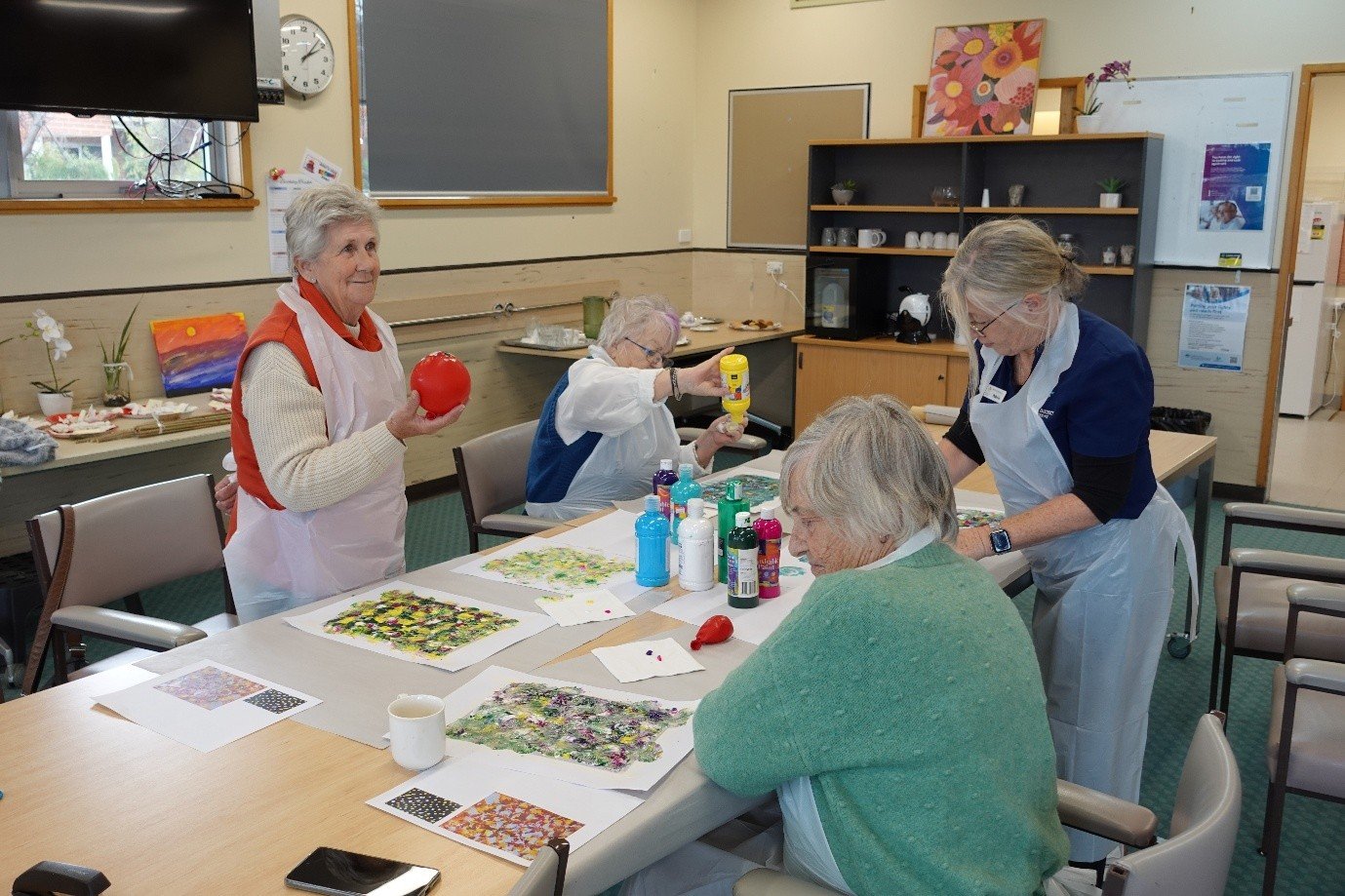 Five elderly women participating in a painting or craft activity in a community room. They are using paints, brushes, and other art supplies on a table. The room has decorations, artwork, and a large clock on the wall.