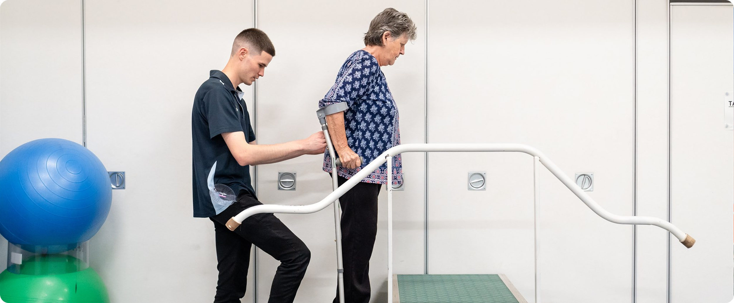 A young man assisting an elderly woman using a crutch to walk during physical therapy, with exercise balls in the background.
