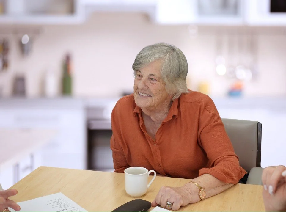 An elderly woman with gray hair, wearing an orange blouse, smiling and sitting at a wooden table with a white mug in front of her in a bright kitchen.