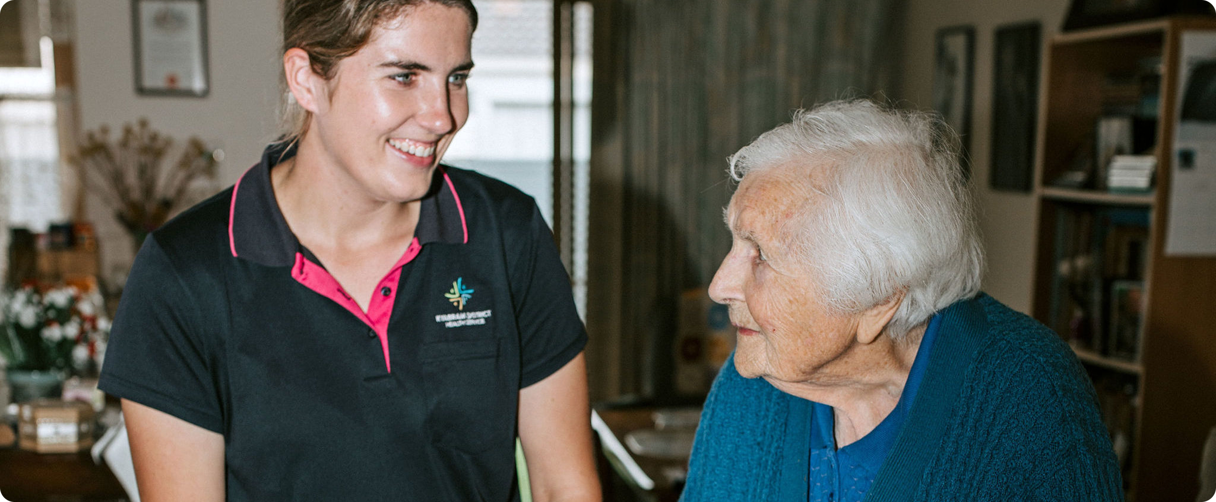 A young woman with short brown hair smiling and talking to an elderly woman with white hair. The young woman is wearing a dark polo shirt with pink accents and a logo, while the elderly woman is wearing a blue sweater.