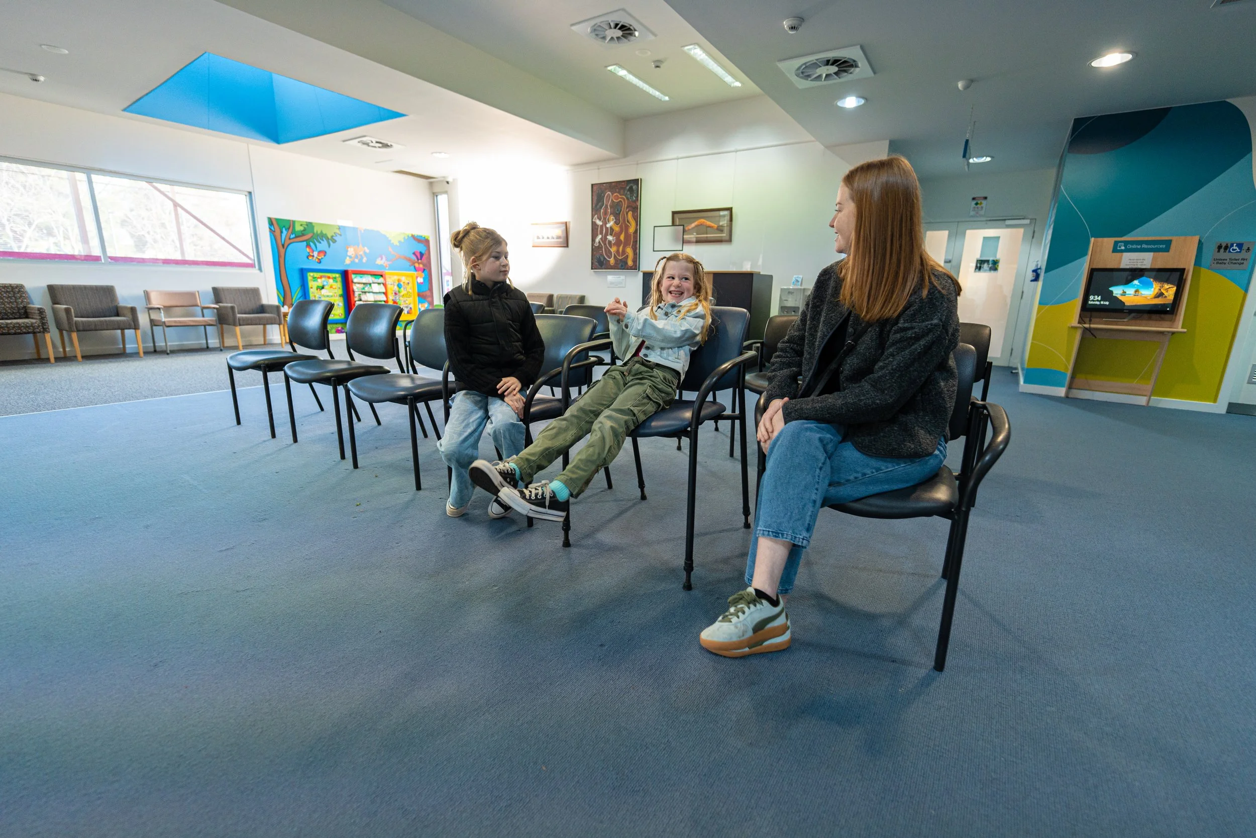A waiting room with chairs along the wall, three women sitting and chatting, one of whom is a woman with red hair and two young girls, all smiling.