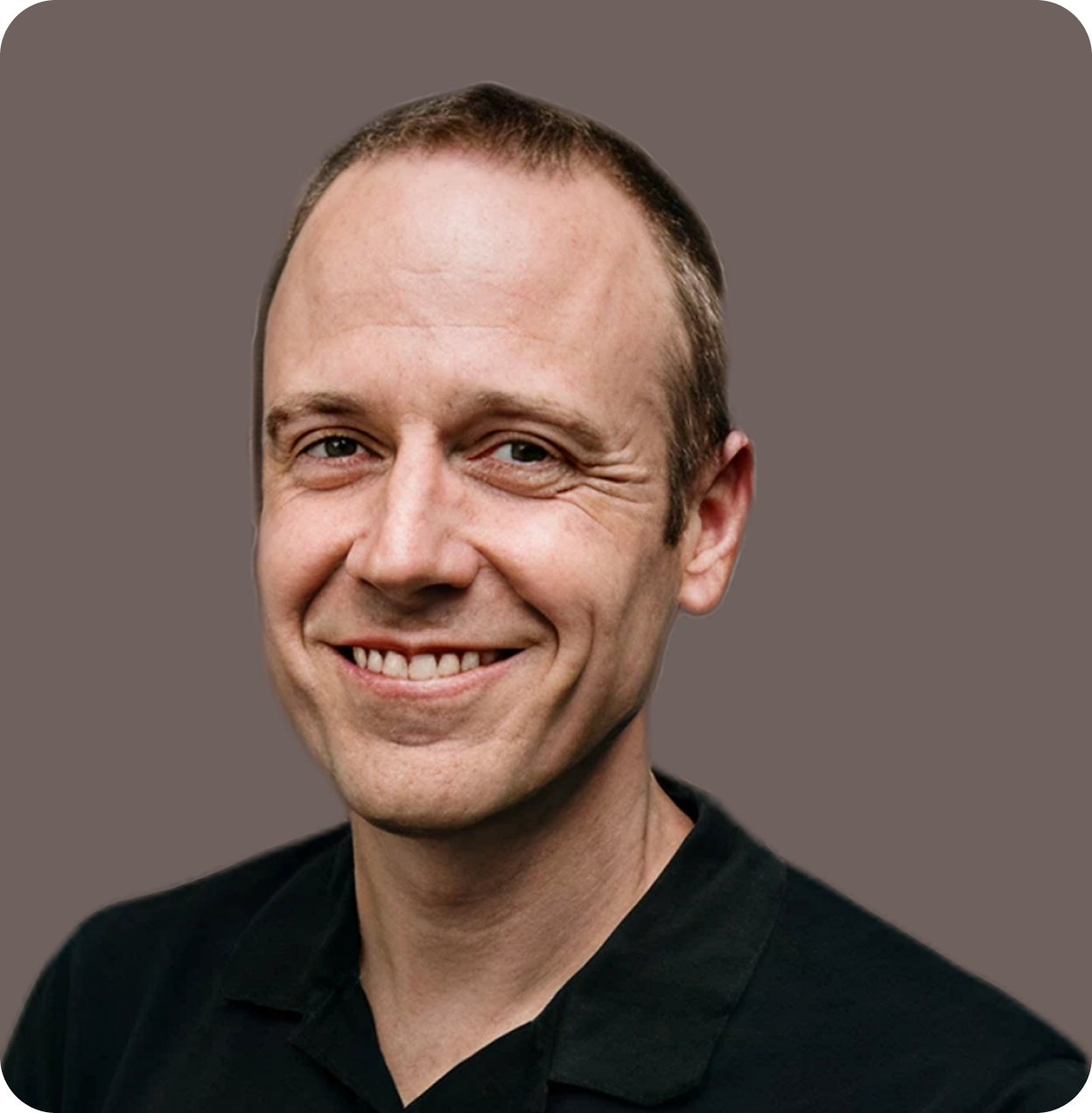 A smiling man with short brown hair, wearing a black shirt, against a plain gray background.