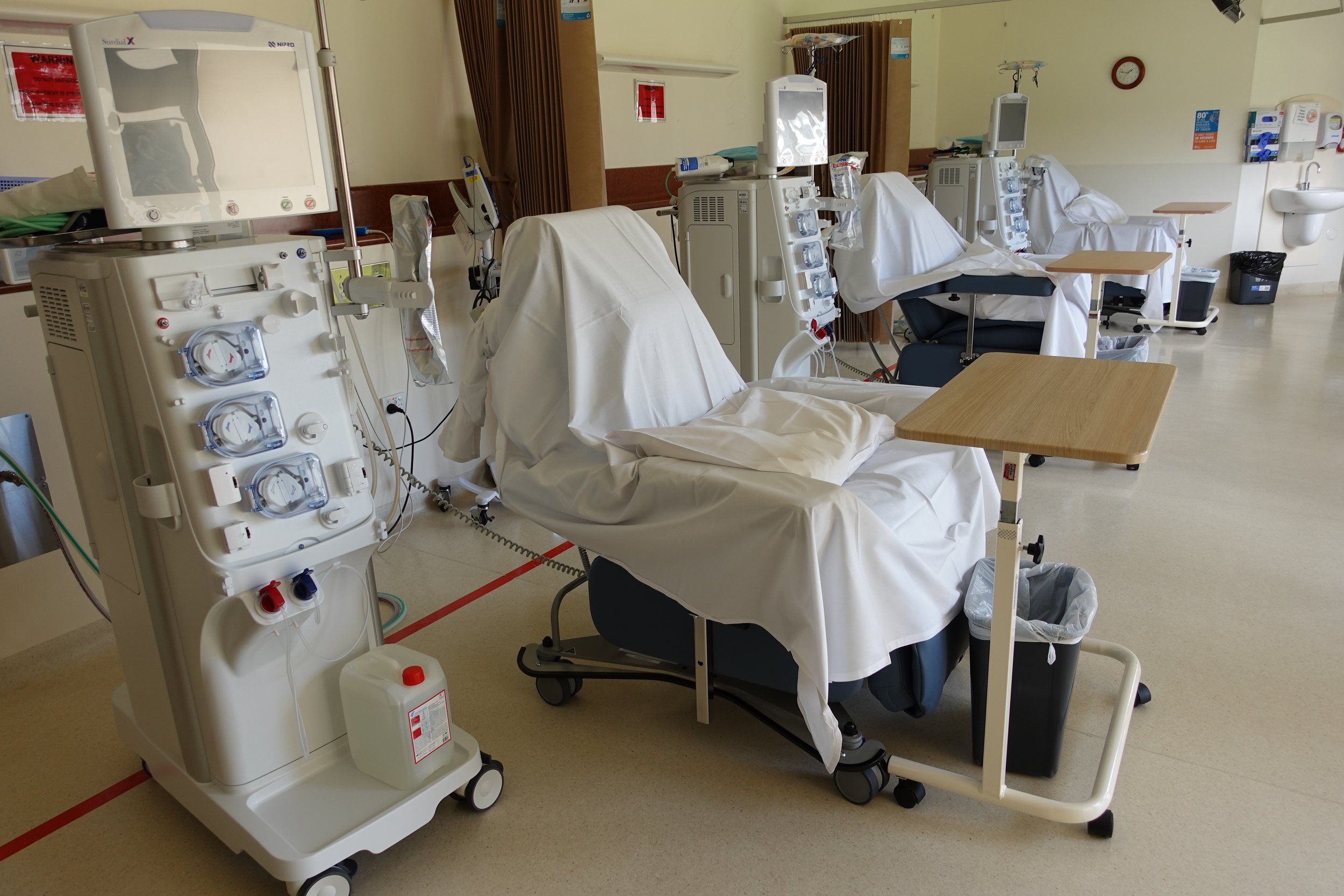 Medical dialysis station with beds covered in white sheets, dialysis machines, and medical equipment in a hospital room.