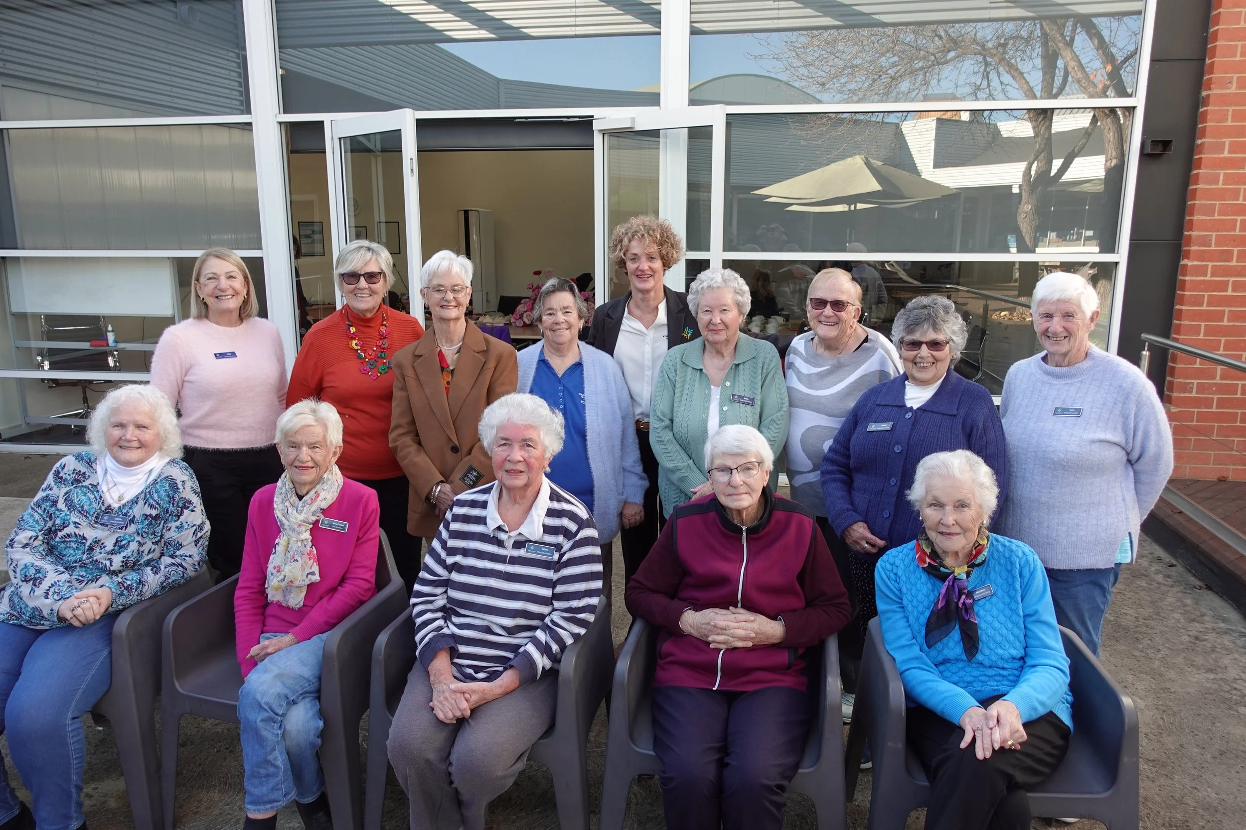 Group of elderly women and caregivers posing for a photo outside a building with large windows and glass door, some seated and some standing, smiling.