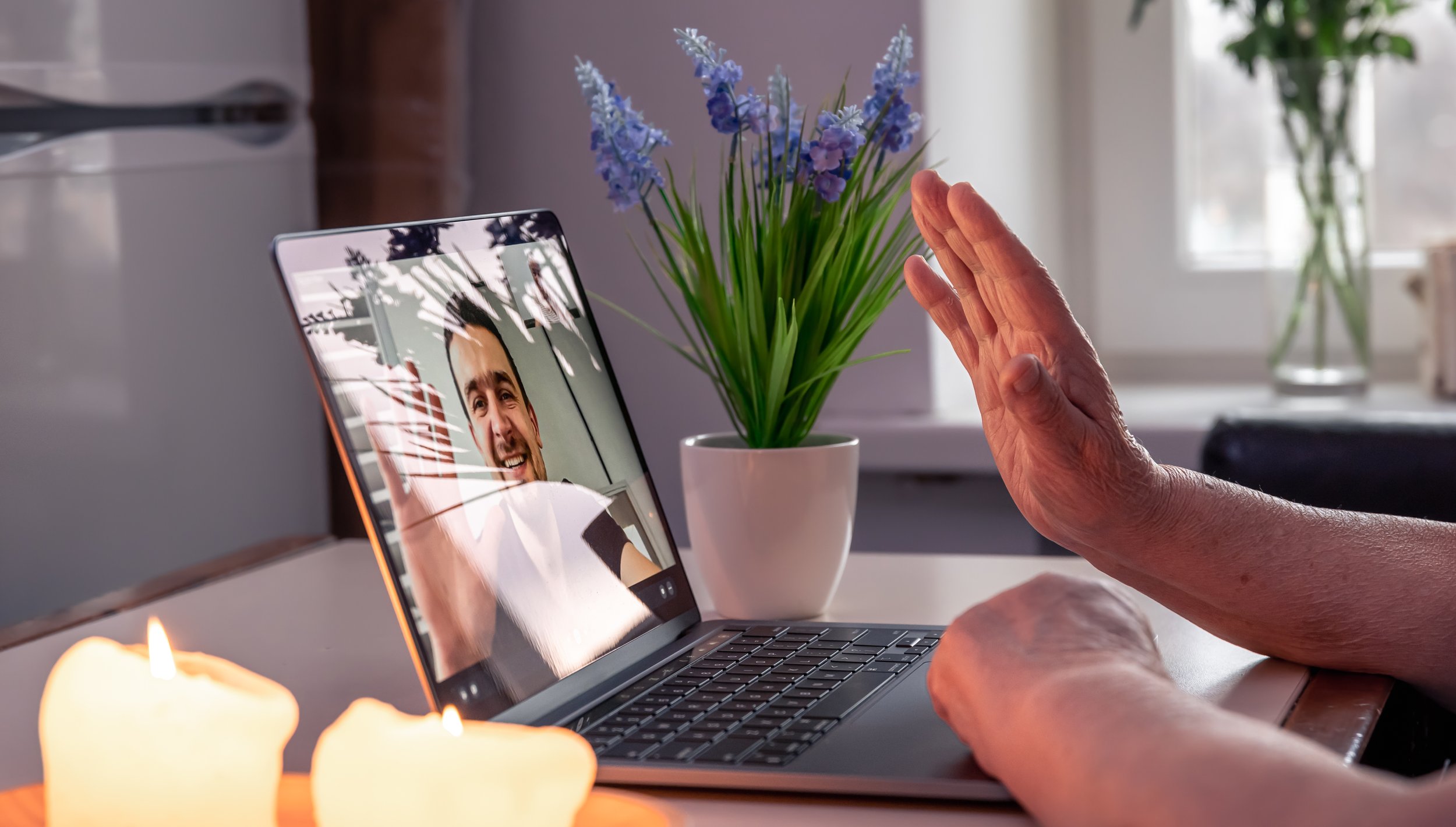 A person waving at a laptop screen displaying a video call with a smiling man. There is a potted plant with purple flowers on the desk and some lit candles in the foreground.