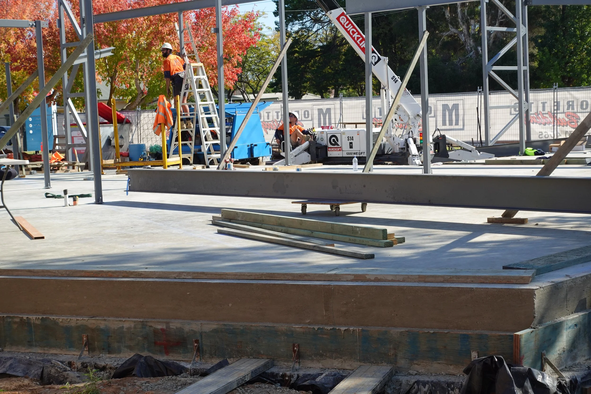 Construction workers building a steel frame structure outdoors with trees and autumn foliage in the background.