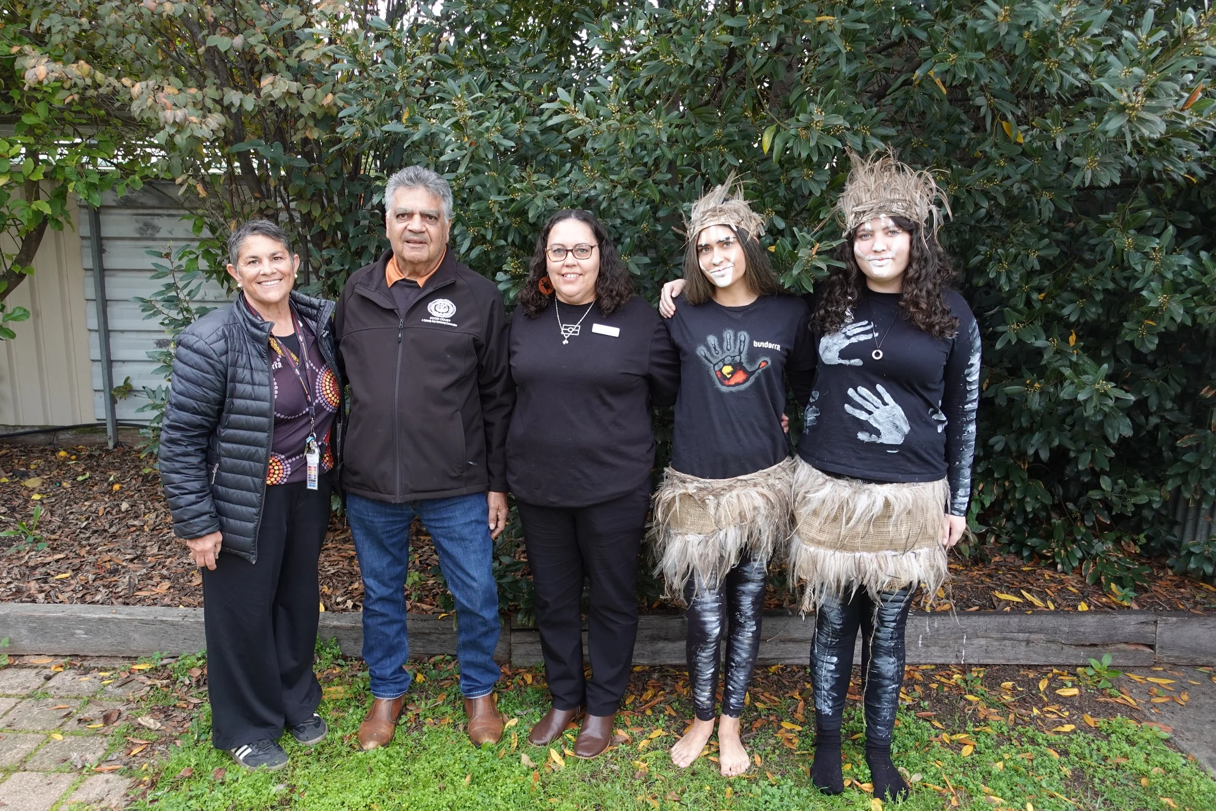 Five people standing outdoors in front of dense green bushes. Two women on right are dressed in traditional or cultural costumes with face paint and grass skirts, while the others are in casual or professional attire. All are smiling or posing casually.