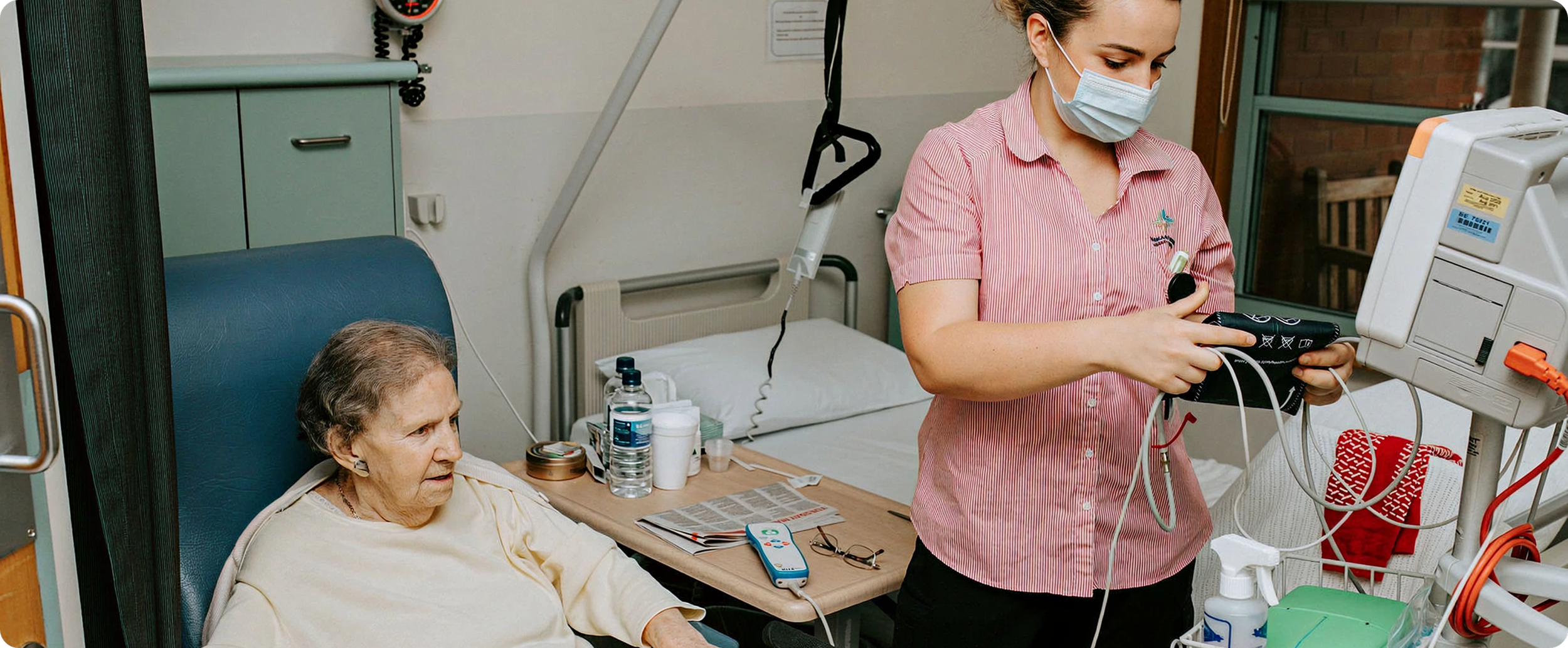 A nurse in a pink uniform and face mask checks a blood pressure monitor on a senior woman patient in a hospital room.