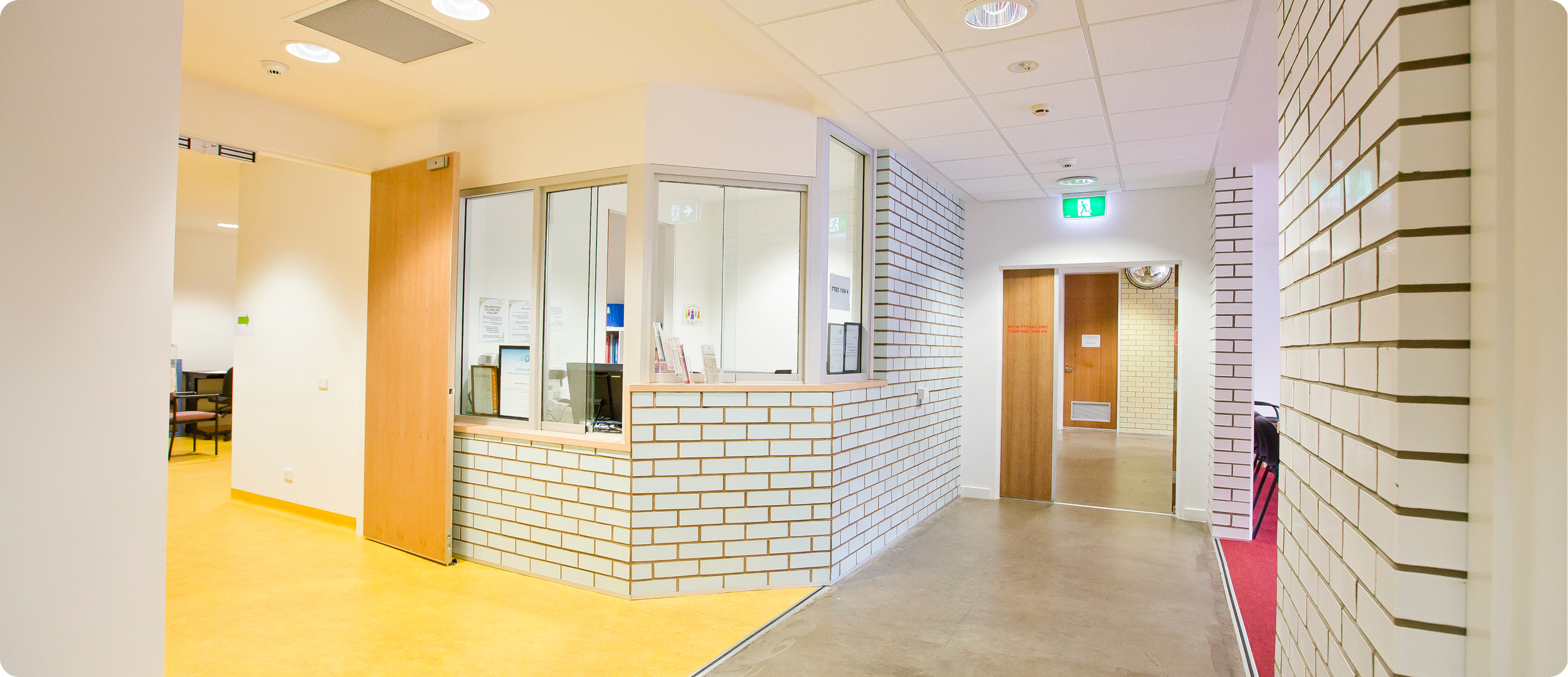 A reception area in a healthcare or office building with white brick walls, a wooden counter with computers, and a hallway with wooden doors and an emergency exit sign.