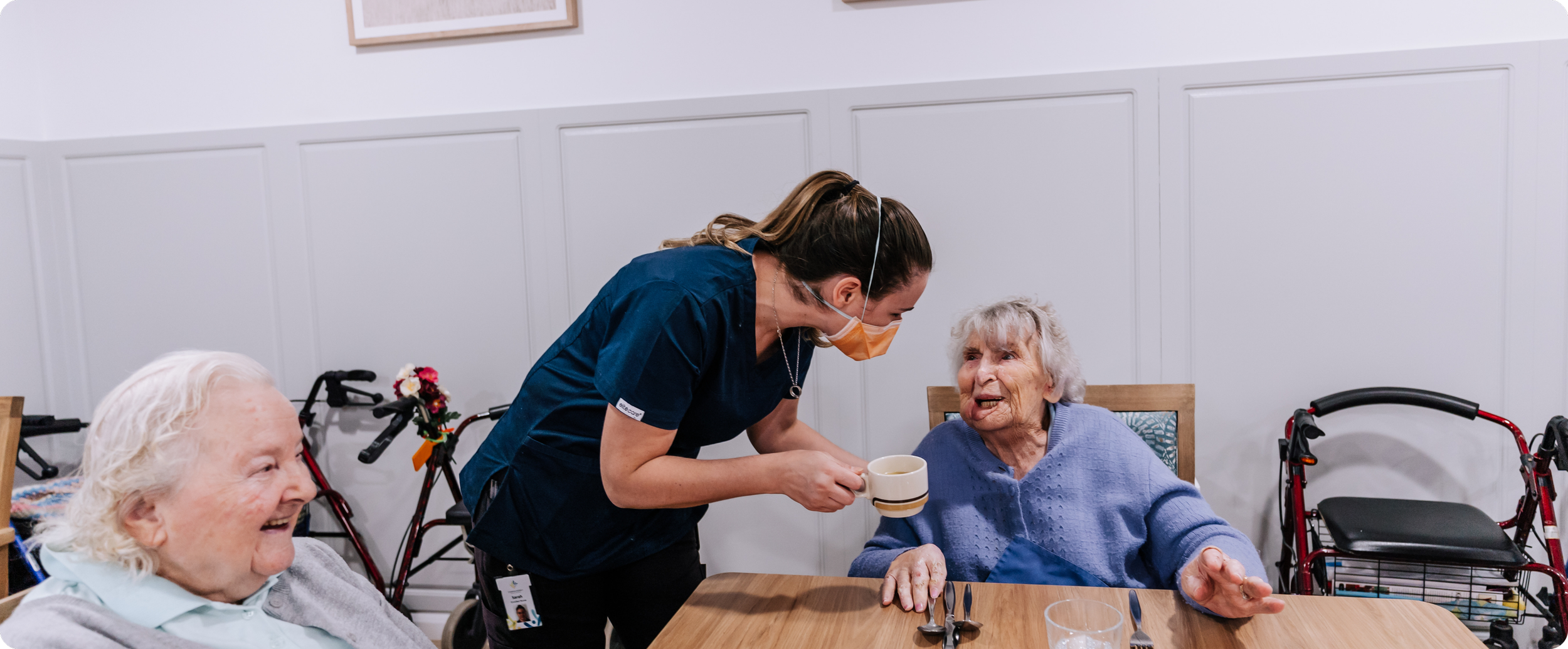 A caregiver with a face mask handing a cup to an elderly woman with white hair, seated at a dining table in a care facility. Two other elderly women are seated nearby, one smiling and the other with a walker nearby.