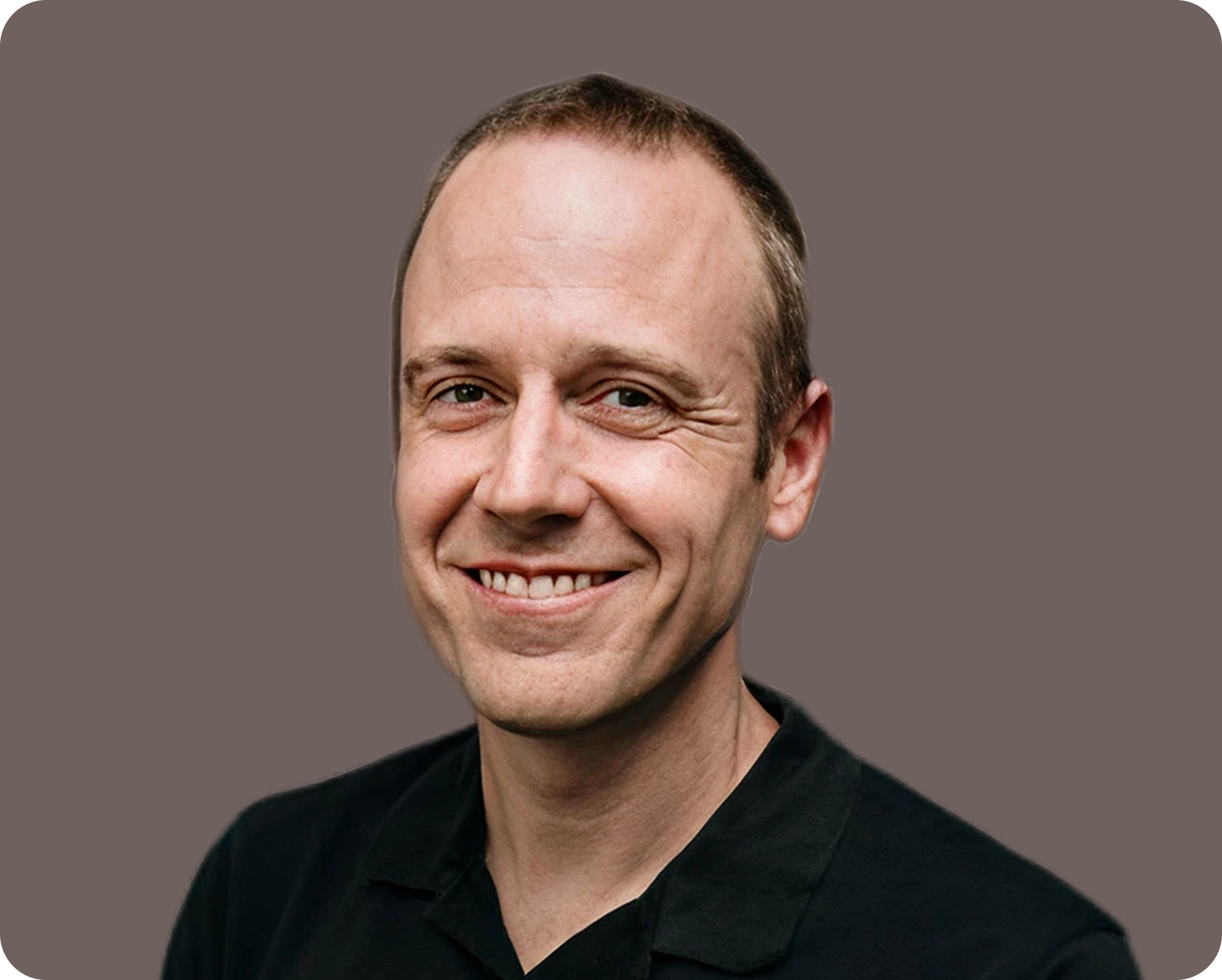 A smiling man with short hair and wearing a black shirt, against a plain background.