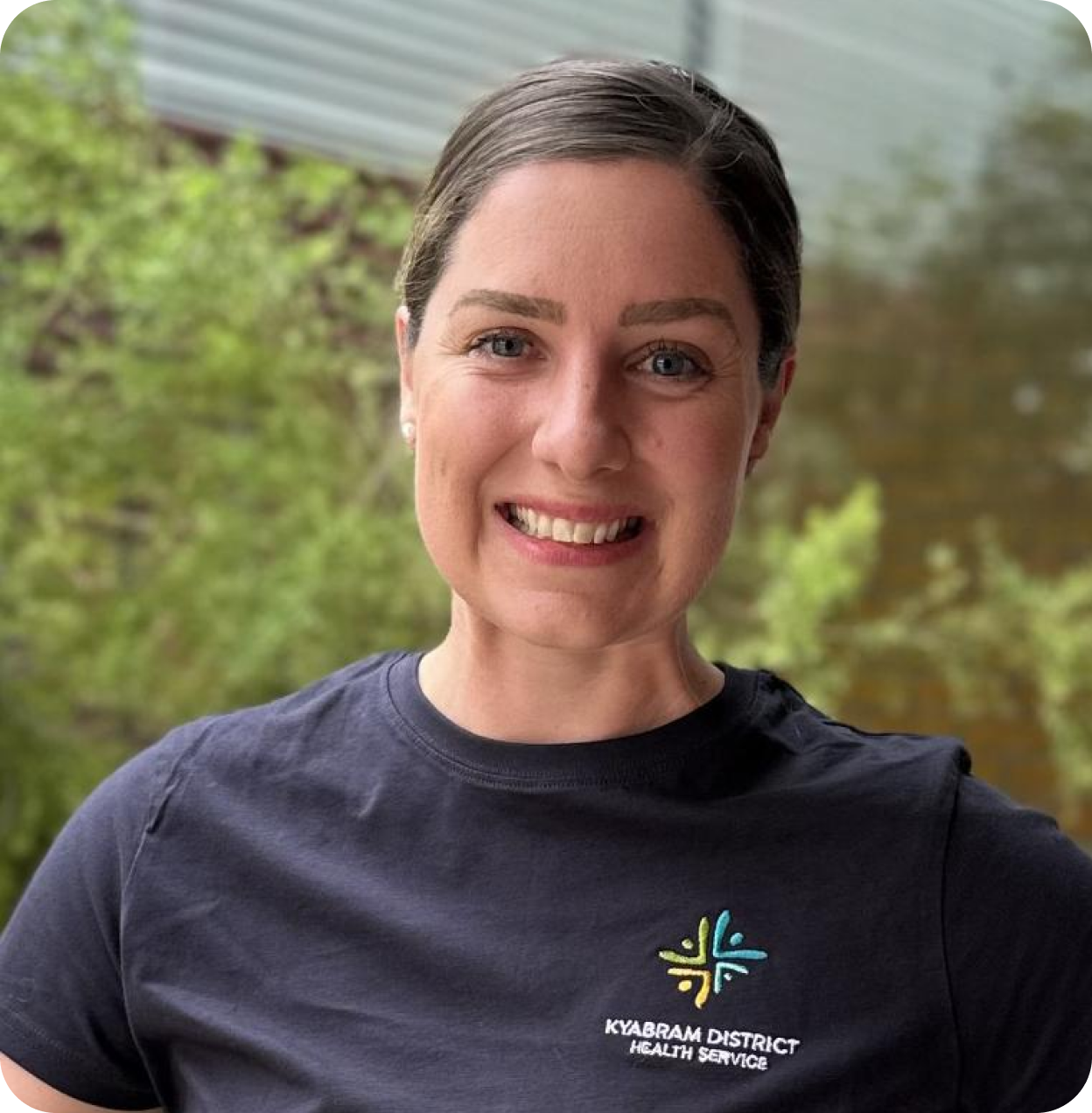 A woman smiling outdoors, wearing a navy T-shirt with 'Kyabram District Health Service' logo, standing in front of green trees and a body of water.