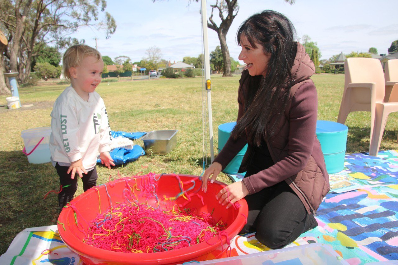 A woman and a young girl playing with colorful rubber bands and yarn in a large red basin outdoors, on a grassy field with trees and chairs in the background.