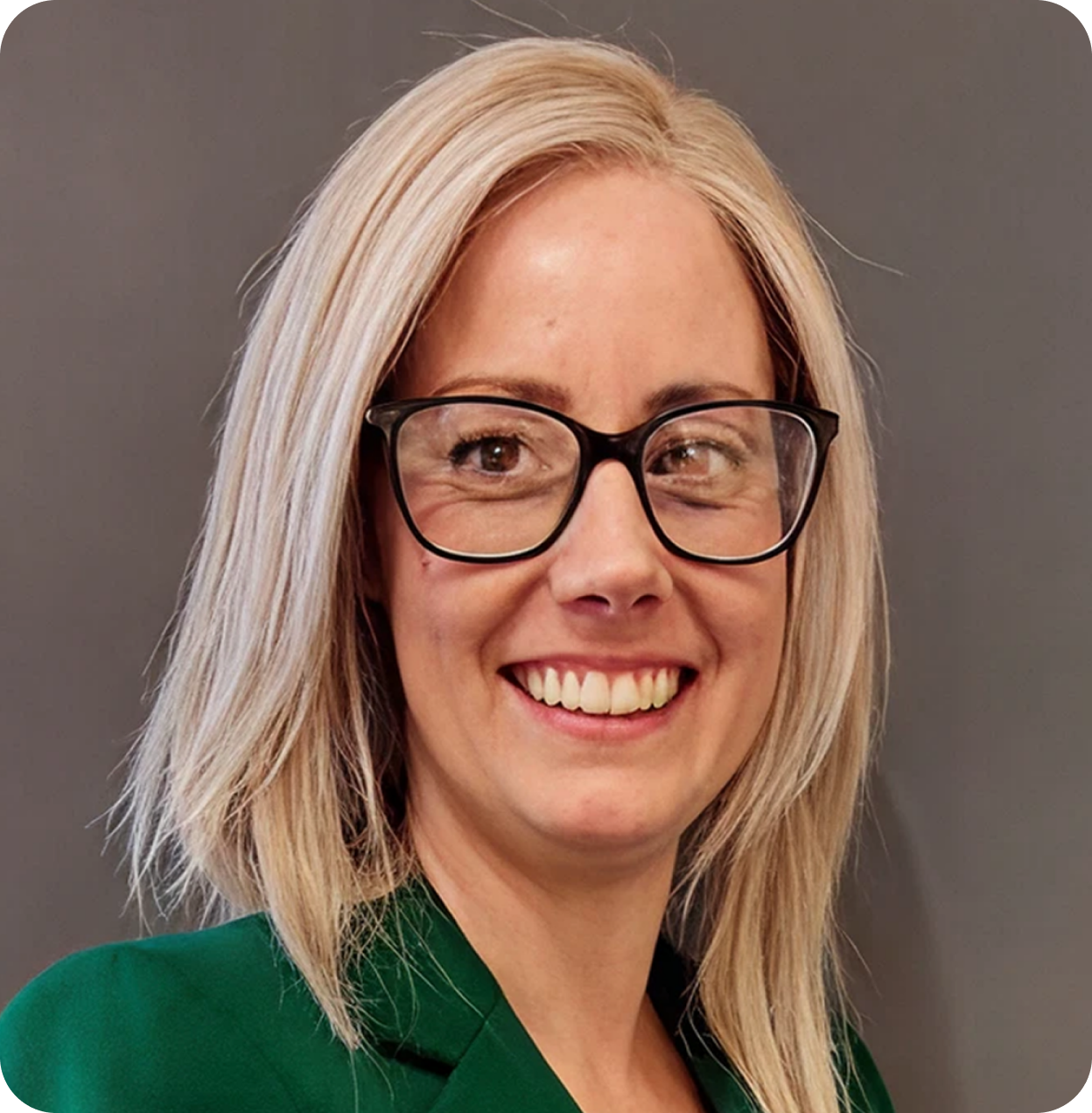 A woman with blonde hair, wearing glasses, smiling, and dressed in a green blazer against a plain background.