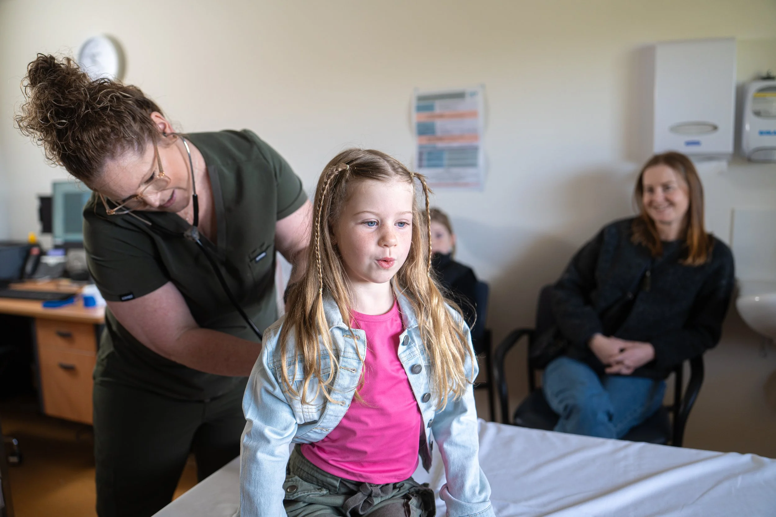 A young girl with braided hair positioned on a hospital bed, being attended to by a female healthcare professional, while a woman and other children are seated in the background in a medical office.
