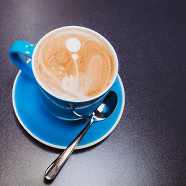 A blue coffee cup filled with frothy coffee on a matching blue saucer, with a small silver spoon resting on the saucer, all on a dark surface.