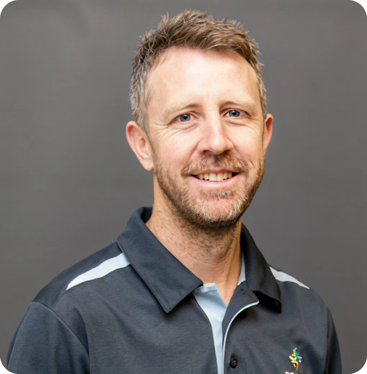 A smiling man with short light brown hair, blue eyes, and a beard, wearing a black sports shirt with white accents and a small colorful logo on the chest, standing against a plain gray background.