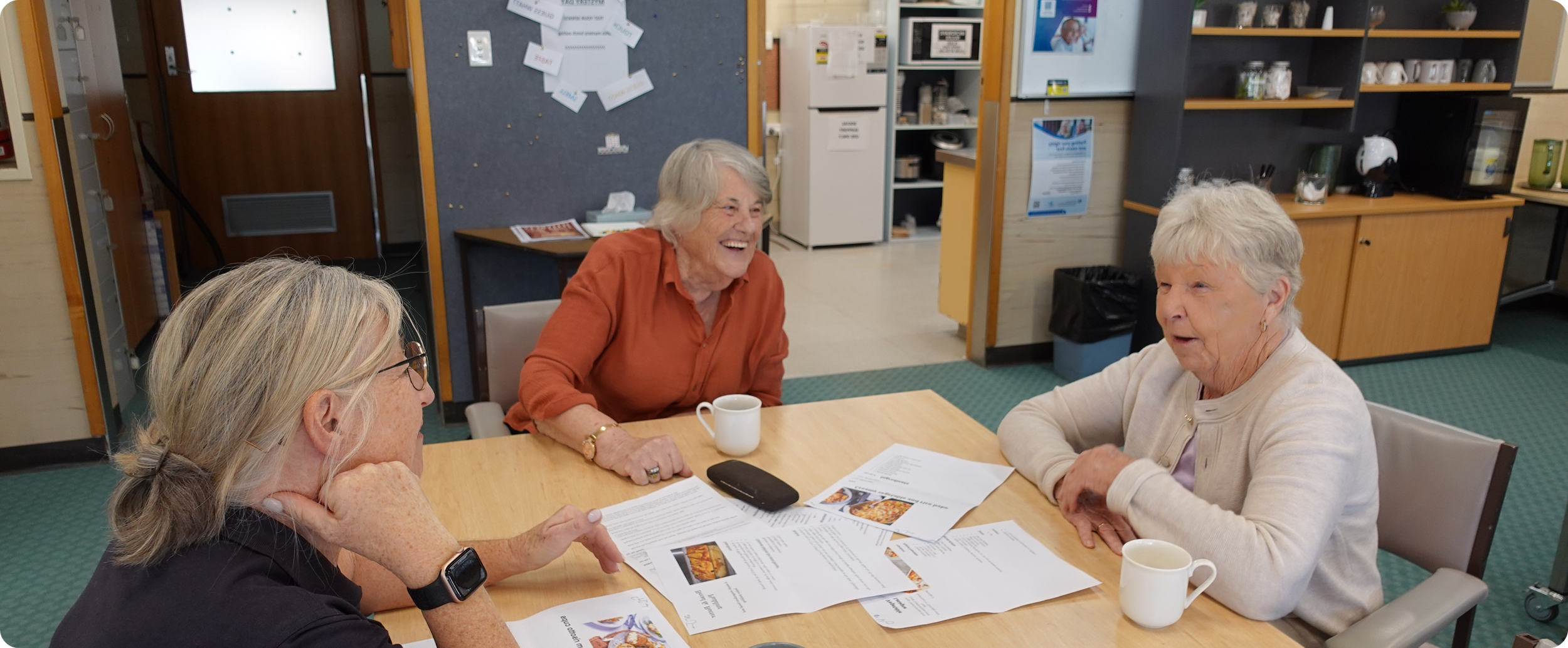 Three elderly women sitting at a table, engaging in a conversation with papers and menus in front of them, in a casual meeting room.